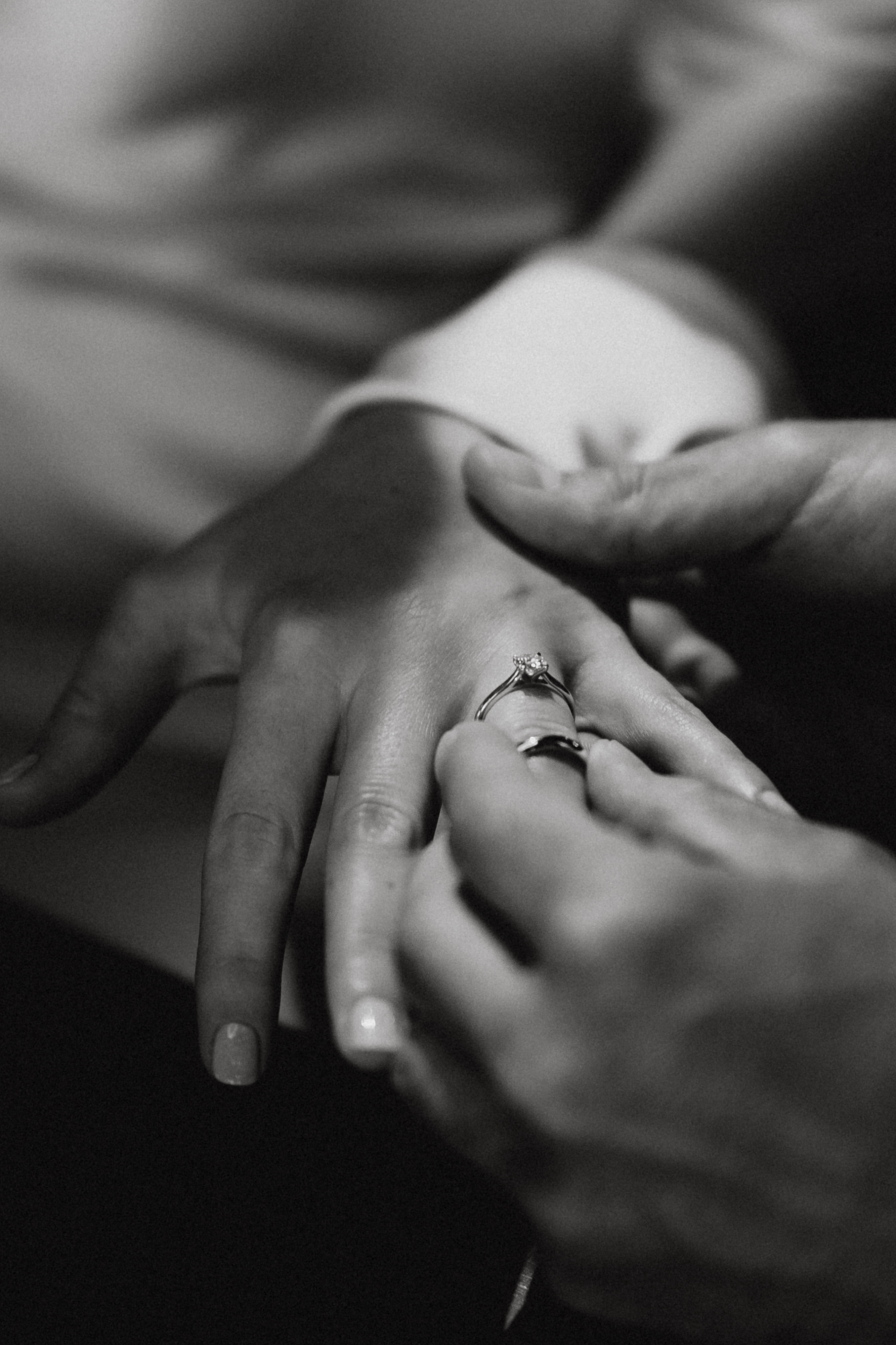 Schwarzweißfoto einer Person, die einer anderen Person einen Ring an den Finger steckt. Der Fokus liegt auf den Händen, wodurch der Ring hervorgehoben wird, der gerade angesteckt wird. Aufgenommen von der talentierten Fotografin Tasha Ender während eines professionellen Fotoshootings. Der Hintergrund ist leicht verschwommen, was den intimen Moment betont.