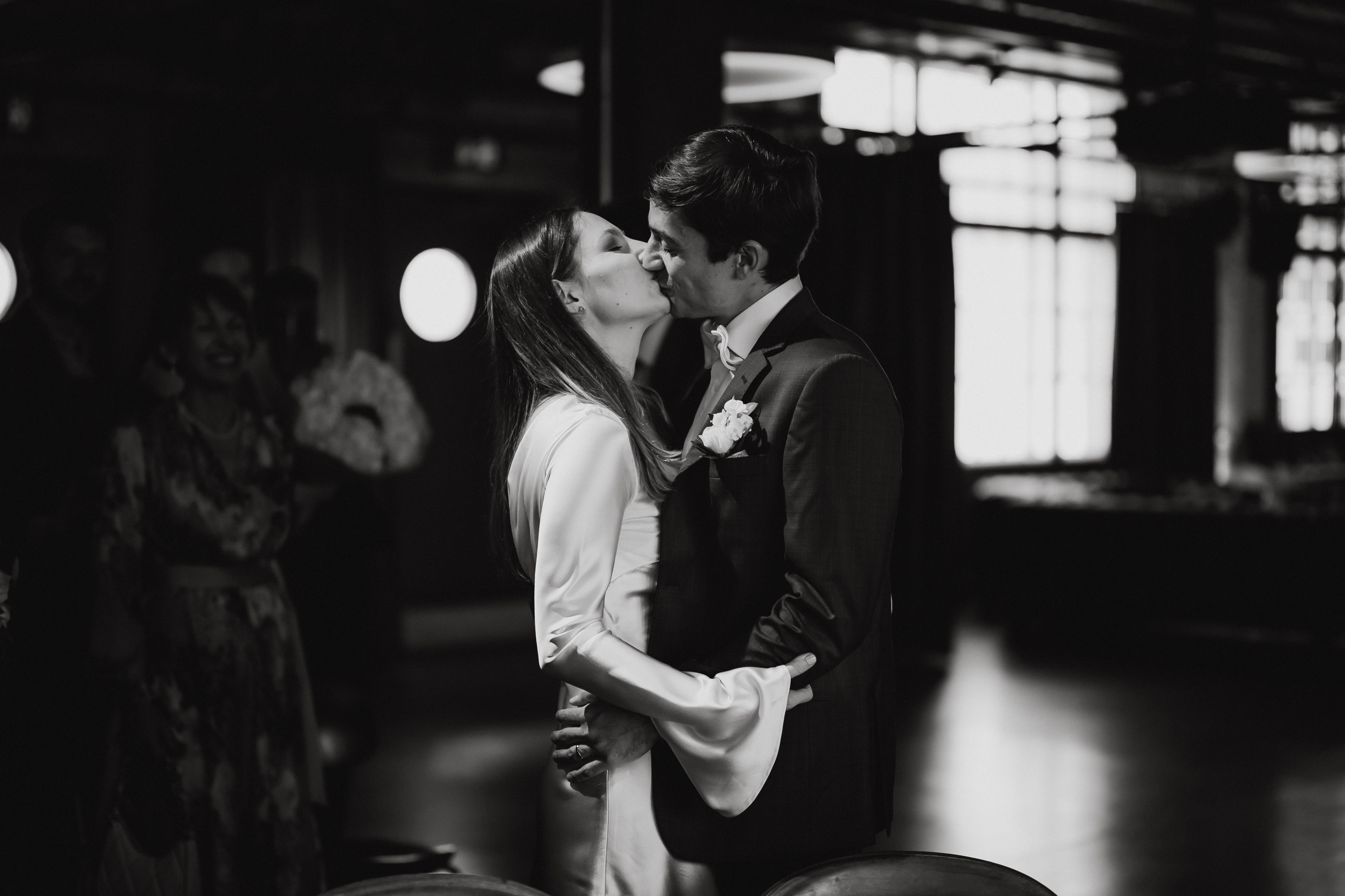 Ein Schwarzweißfoto eines talentierten Fotografen zeigt ein Paar in Abendgarderobe, das sich in einem warm beleuchteten Innenraum küsst. Die Frau im weißen Kleid hält die Schultern des Mannes, während er im Anzug mit Ansteckblume seine Arme um ihre Taille legt. Im Hintergrund sind verschwommene Figuren zu sehen.