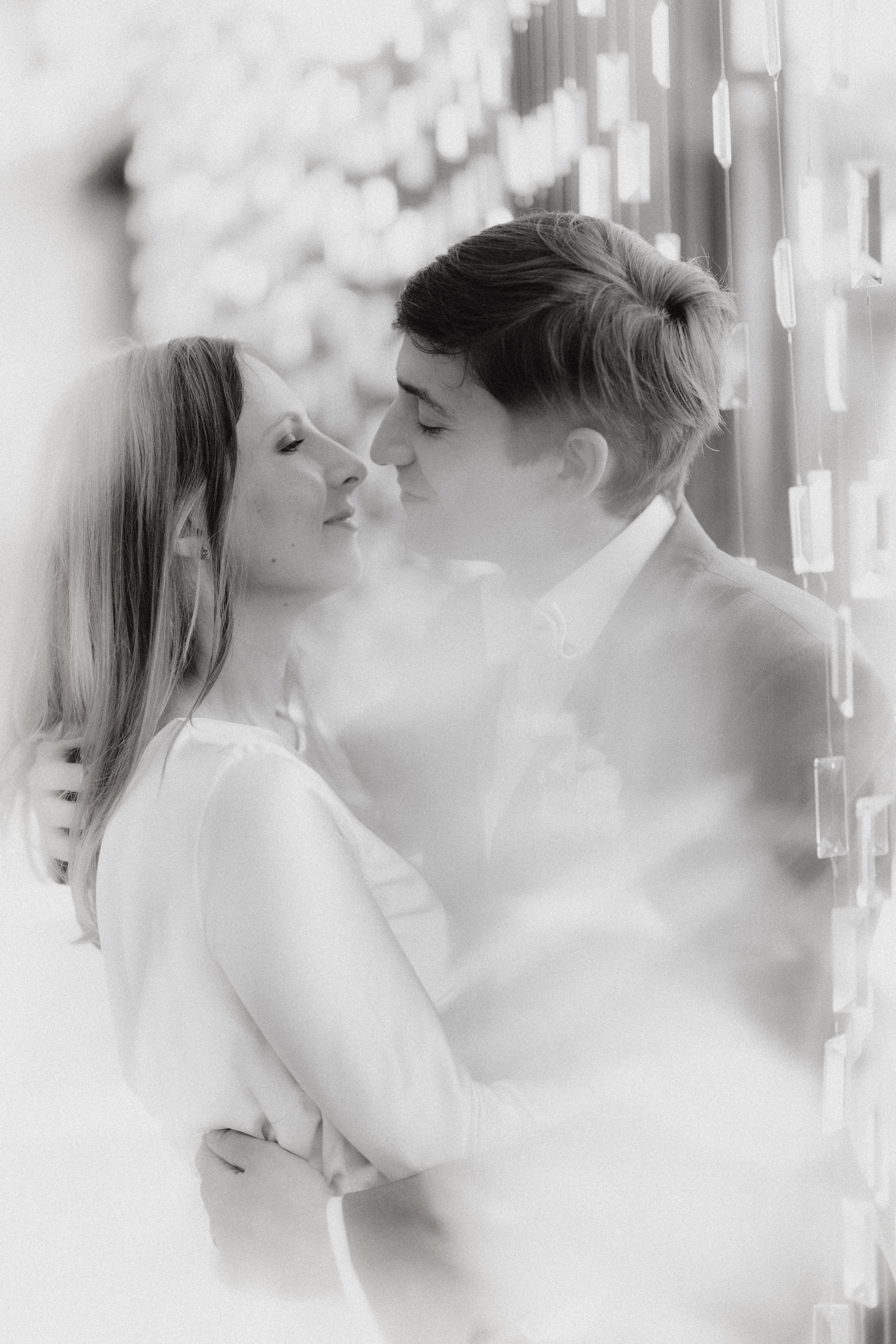 Ein Schwarzweißfoto von Tasha Ender, einer renommierten Fotografin von Fotograf Baden, zeigt ein Paar, das sich umarmt und liebevoll anlächelt. Die Frau links hat lange Haare und trägt ein helles Oberteil. Der Mann rechts hat kurze Haare und trägt einen Anzug. Weiches Licht und Hintergrundelemente erzeugen einen verträumten Effekt.