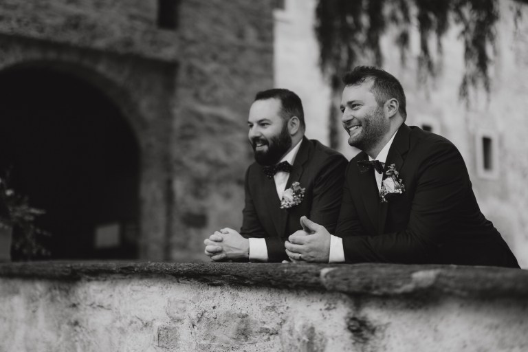 Eine zeitlose und ruhige Atmosphäre ziert die Natur in Zürich, wo zwei Männer in Anzügen an einer Steinmauer lehnen, lächeln und in die Ferne blicken. Beide sind mit Fliegen und Boutonnieren geschmückt, was auf einen formellen Anlass schließen lässt. Das rustikale Steingebäude und das umgebende Grün bilden die perfekte Kulisse für die Feier ihrer LGBTQ-Liebe, wunderschön eingefangen durch die Hochzeitsfotografie-Dienste von Tasha Ender Photography.