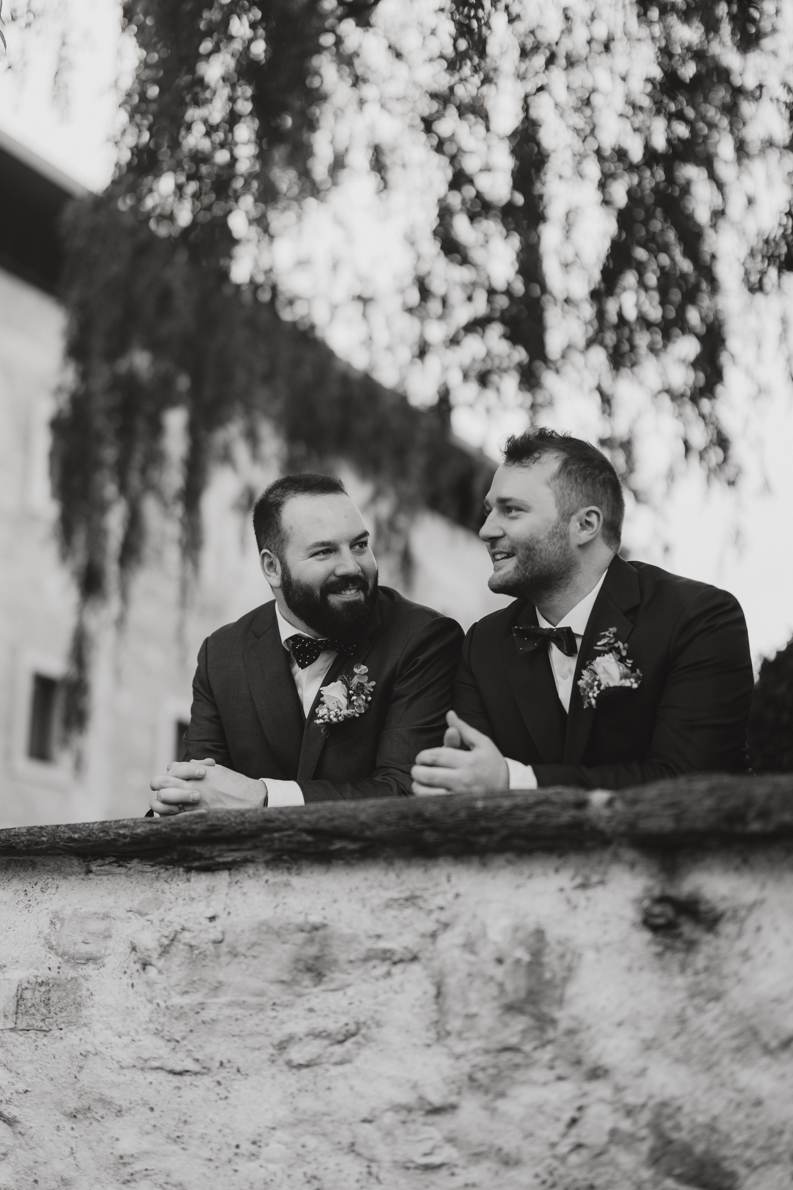 Schwarz-Weiß-Bild vom Hochzeitsfotografie-Service von Tasha Ender Photography. Es zeigt zwei Männer in Anzügen, beide mit Boutonnieren geschmückt, die an einer Steinmauer lehnen. Der bärtige Mann links und sein Begleiter lächeln sich herzlich an. Der nostalgische Hintergrund umfasst einen Baum und ein Gebäude, die den Charme des alten Zürich oder vielleicht Baden im Aargau heraufbeschwören.