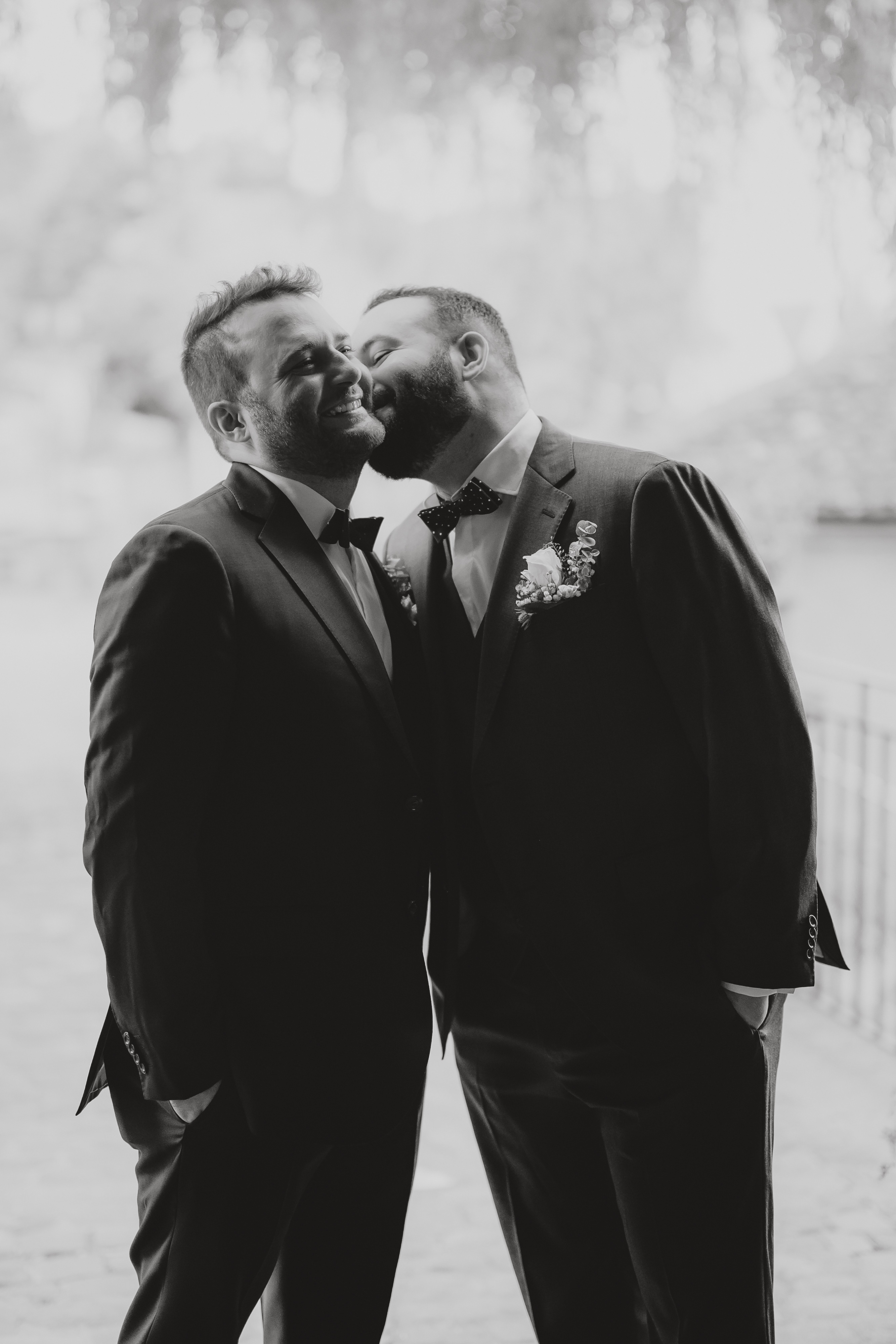 Ein Schwarzweißfoto von Tasha Ender Photography, das zwei Männer in Anzügen zeigt, die eng beieinander stehen und sich herzlich anlächeln. Ein Mann küsst den anderen auf die Wange, während sie Händchen halten, die beide mit Boutonnieren geschmückt sind. Sie scheinen einen freudigen und liebevollen Moment zu teilen, möglicherweise bei einer Hochzeit oder einem offiziellen Anlass in Baden oder an einem anderen Ort im Aargau.