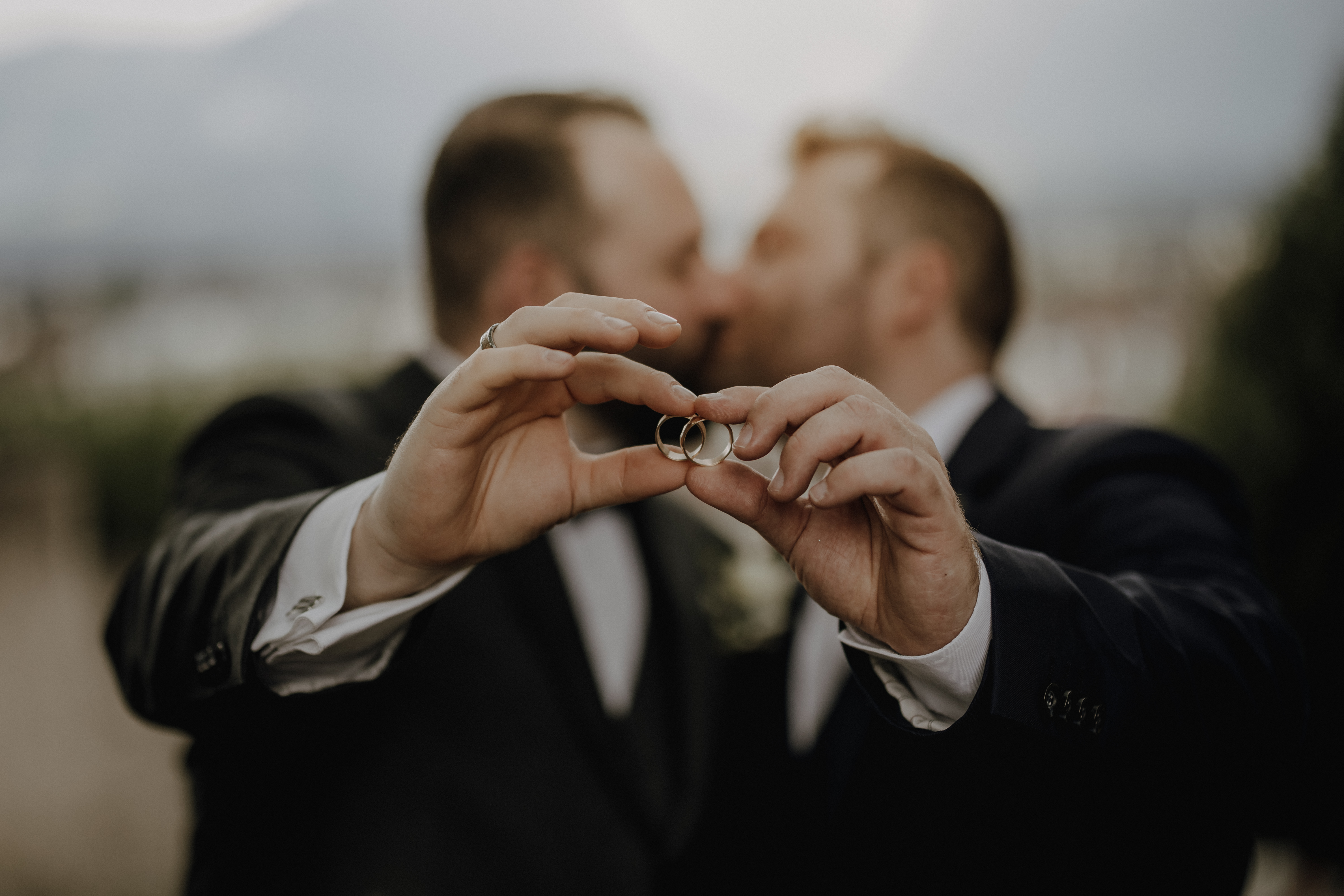 Zwei Männer in formeller Kleidung halten ihre Eheringe zusammen, wunderschön eingefangen von Hochzeitsfotografie von Tasha Ender Photography, während sie sich im Hintergrund küssen. Die Ringe und ihre Hände sind scharf vor einer sanft verschwommenen Außenszene zu erkennen, die die natürliche Schönheit von Zürich heraufbeschwört.