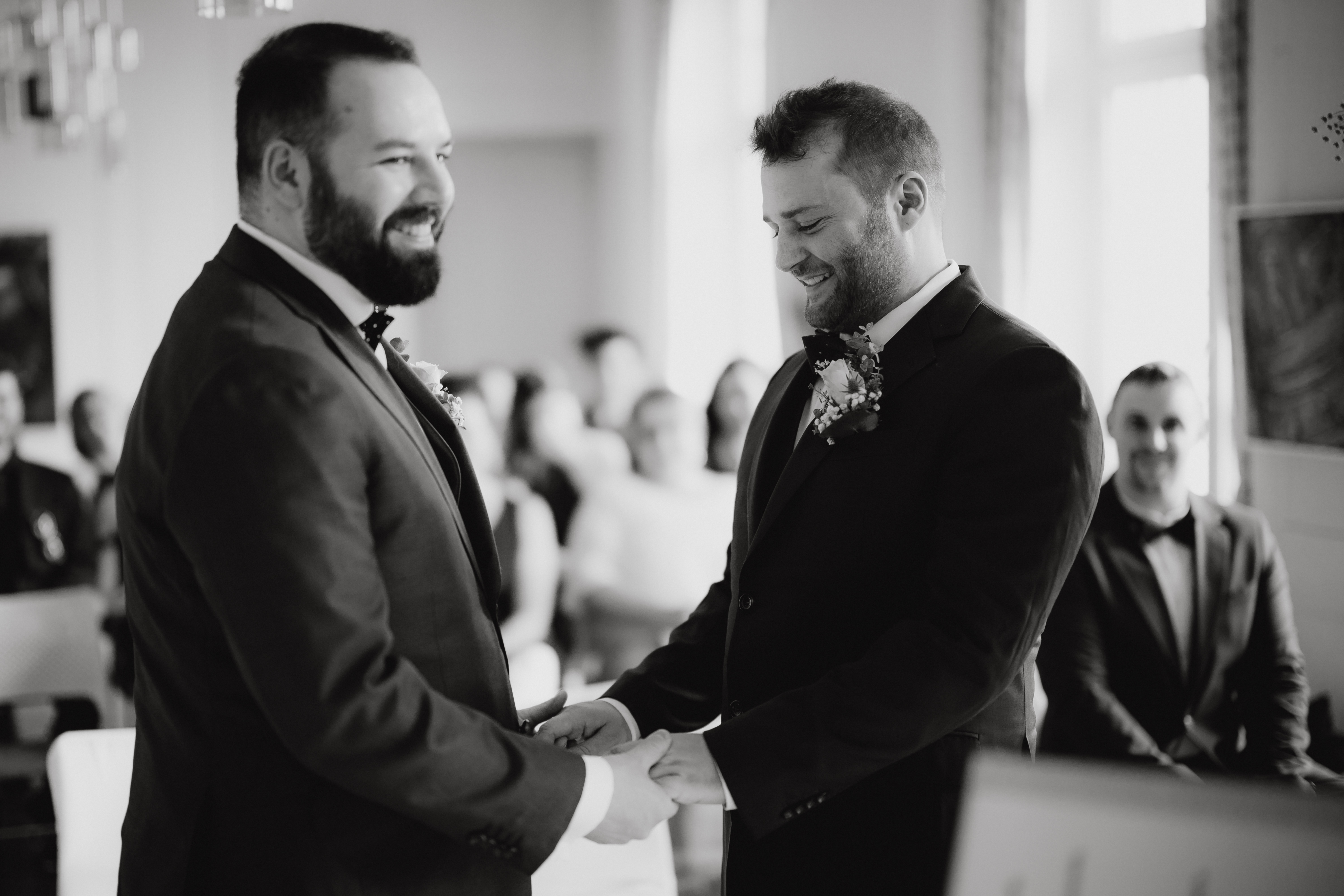 Eine wunderschöne Hochzeitsfotografie von Tasha Ender Photography zeigt ein Schwarzweißfoto von zwei Männern in Anzügen, die während ihrer Hochzeitszeremonie Händchen halten und einander anlächeln, wobei im Hintergrund die sitzenden Gäste sanft unscharf sind.