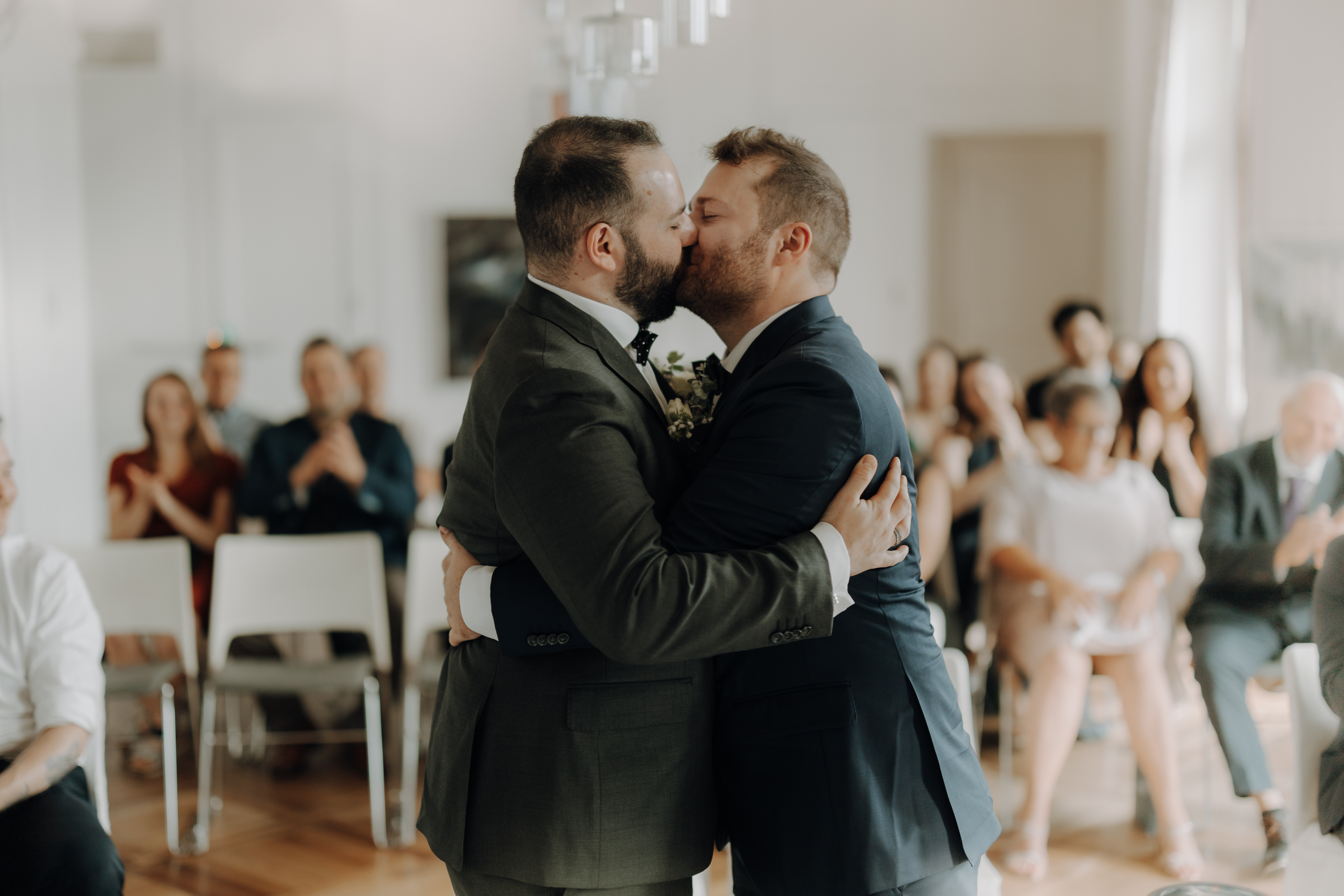 Zwei Männer in Anzügen küssen sich in einem hell erleuchteten Raum, umgeben von einem kleinen Publikum auf weißen Stühlen. Der Moment fängt die Feier ihrer LGBTQ-Hochzeit ein, mit klatschenden und lächelnden Gästen im Hintergrund. Diese berührende Zeremonie fand in Baden, nicht weit von Zürich, statt. Wunderschön festgehalten von *Hochzeitsfotografie* von *Tasha Ender Photography*.