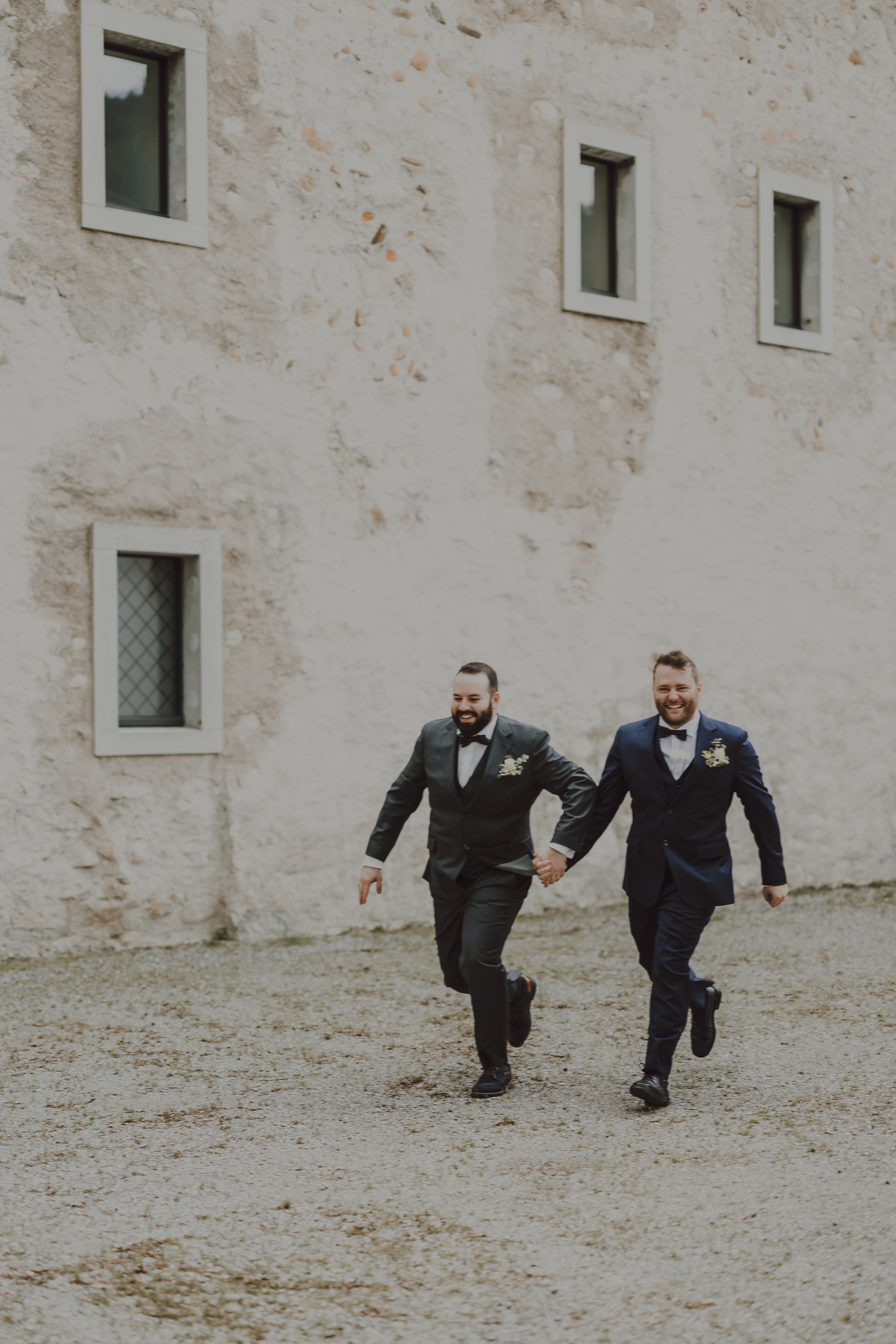 Zwei Männer in formellen Anzügen, Händchen haltend und lächelnd, laufen fröhlich auf einem Kiesweg vor einem rustikalen Gebäude mit kleinen Fenstern. Sie tragen Boutonnieres am Revers, was auf eine festliche, queere Hochzeit schließen lässt. Der Himmel ist bedeckt, was der von ihrer Hochzeitsfotografin eingefangenen Szene einen gedämpften Ton verleiht.