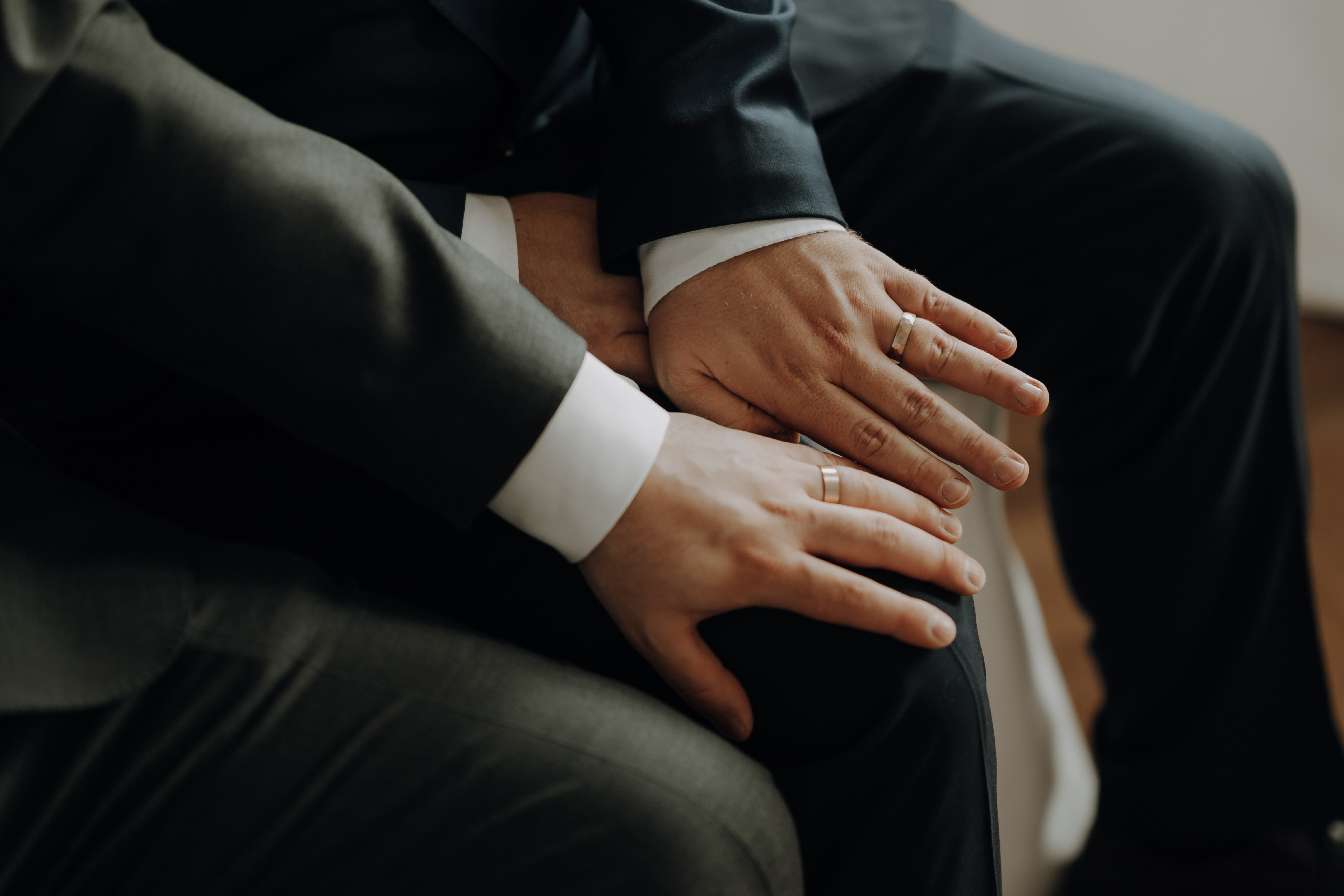 Eine Nahaufnahme aus einer Hochzeitsreportage von Tasha Ender zeigt zwei Menschen in dunklen Anzügen, die eng beieinander sitzen. Ihre Hände ruhen sanft auf den Beinen des jeweils anderen und zeigen ihre Eheringe.