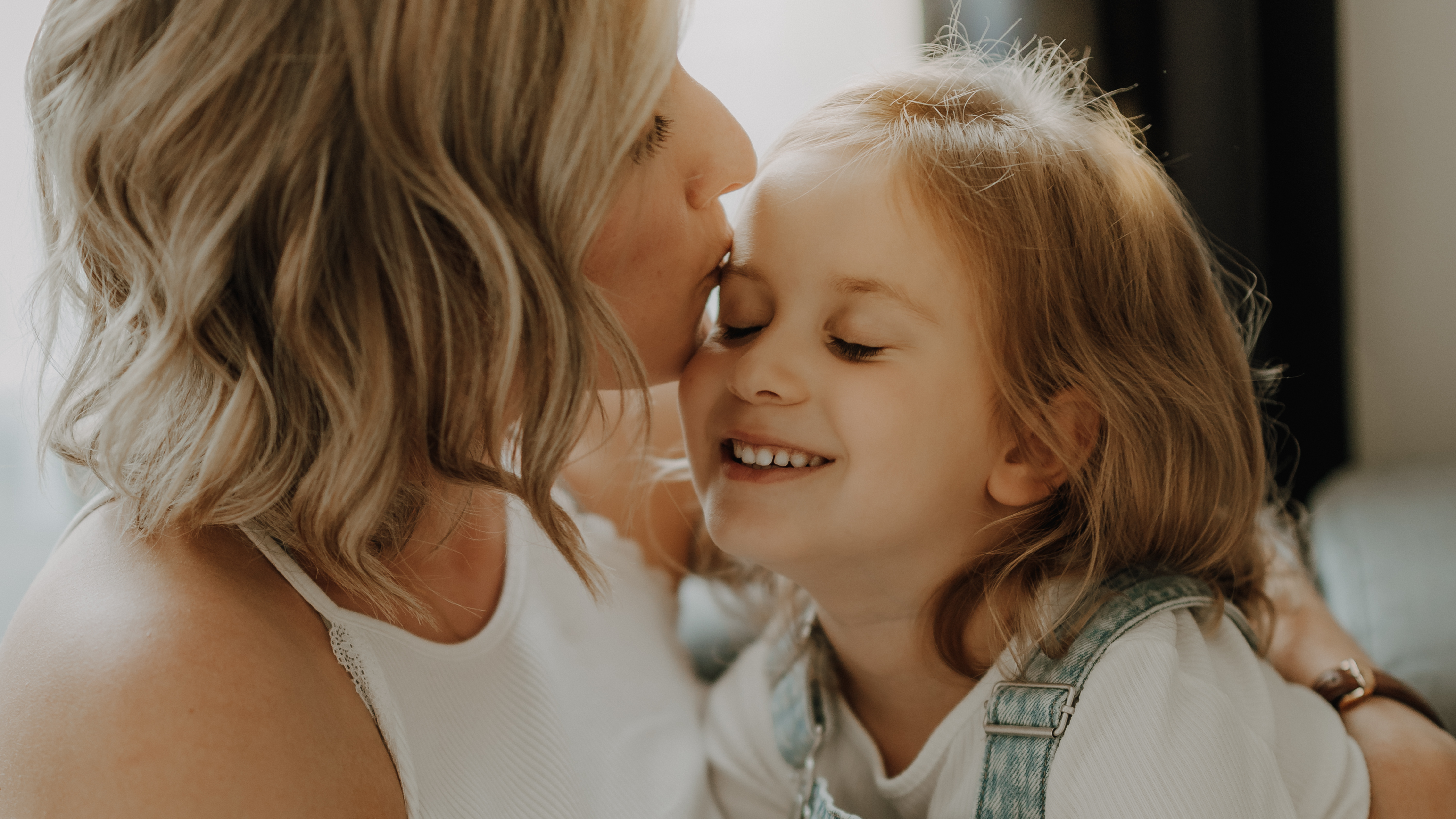 Eine Frau mit kurzen, blonden Haaren küsst ein lächelndes Kind liebevoll auf die Wange. Das Kind, das schulterlanges hellbraunes Haar hat, trägt ein weißes Hemd und einen Jeans-Overall und wirkt auf den natürlichen Familienfotos glücklich und zufrieden.