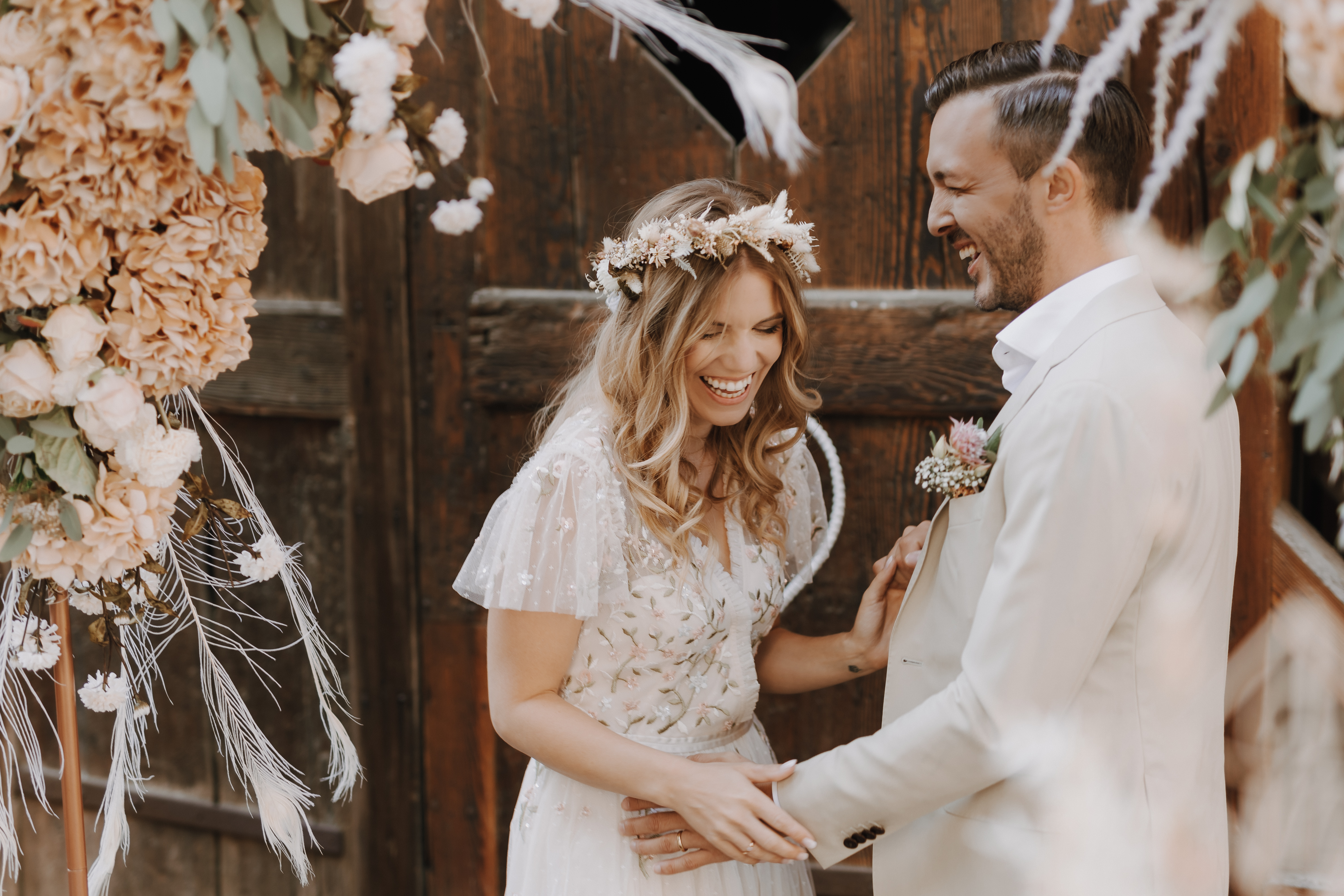 Braut und Bräutigam teilen einen freudigen Moment während ihrer badischen Trauung und lachen zusammen. Die Braut, Tasha Ender, hat lockere Wellen, eine Blumenkrone und ein zartes Spitzenkleid mit kurzen Ärmeln. Der Bräutigam trägt einen hellen Anzug und eine Boutonnière. Sie stehen vor einer rustikalen Holztür, umgeben von Blumenarrangements.