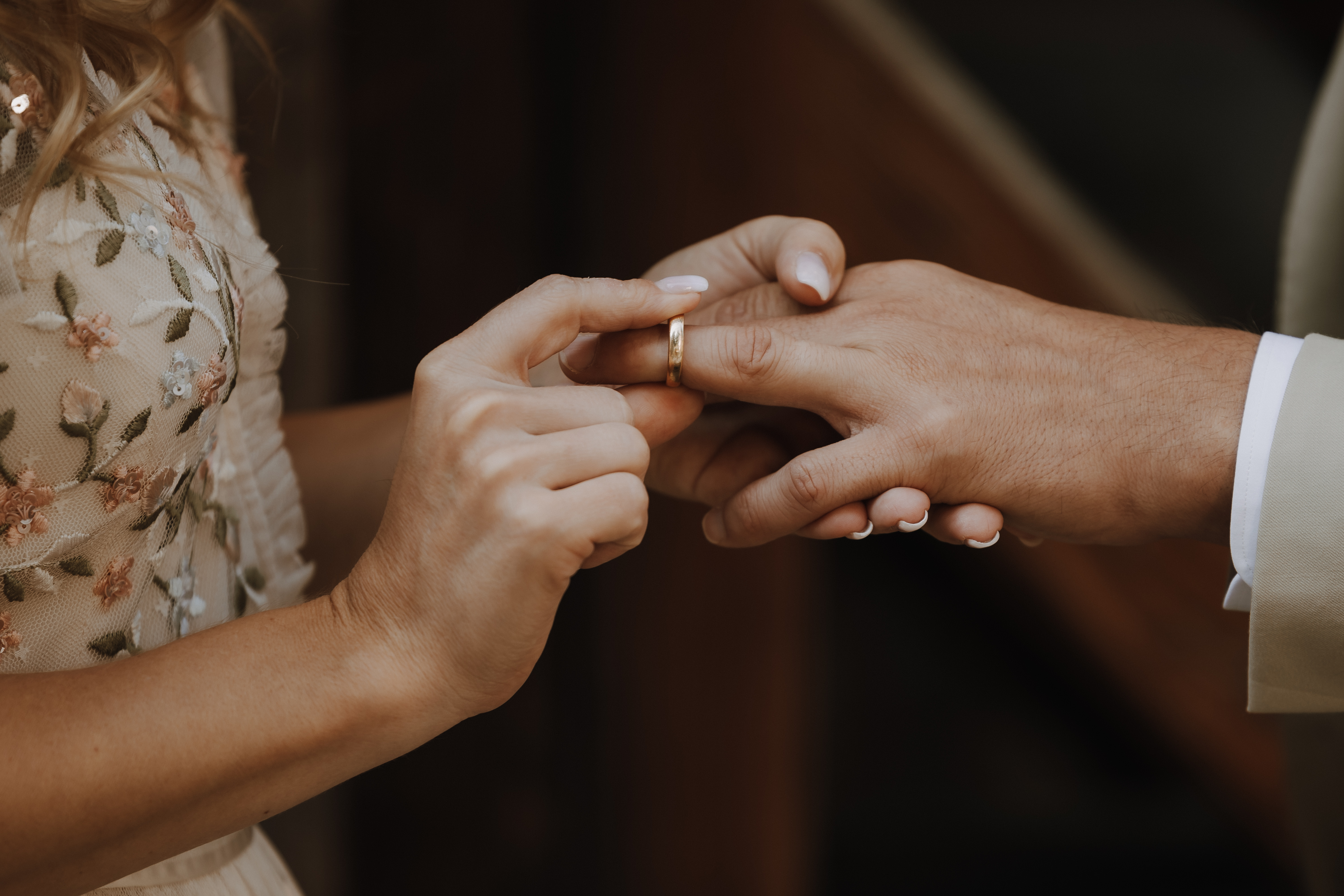 Eine Person in einem beigefarbenen Blumenkleid steckt einer anderen Person einen goldenen Ring an die Hand, was eine Hochzeit oder Verlobung symbolisiert. Der Fokus liegt auf ihren Händen vor einem sanft verschwommenen Hintergrund, wunderschön eingefangen von Hochzeitsfotografin Tasha Ender.