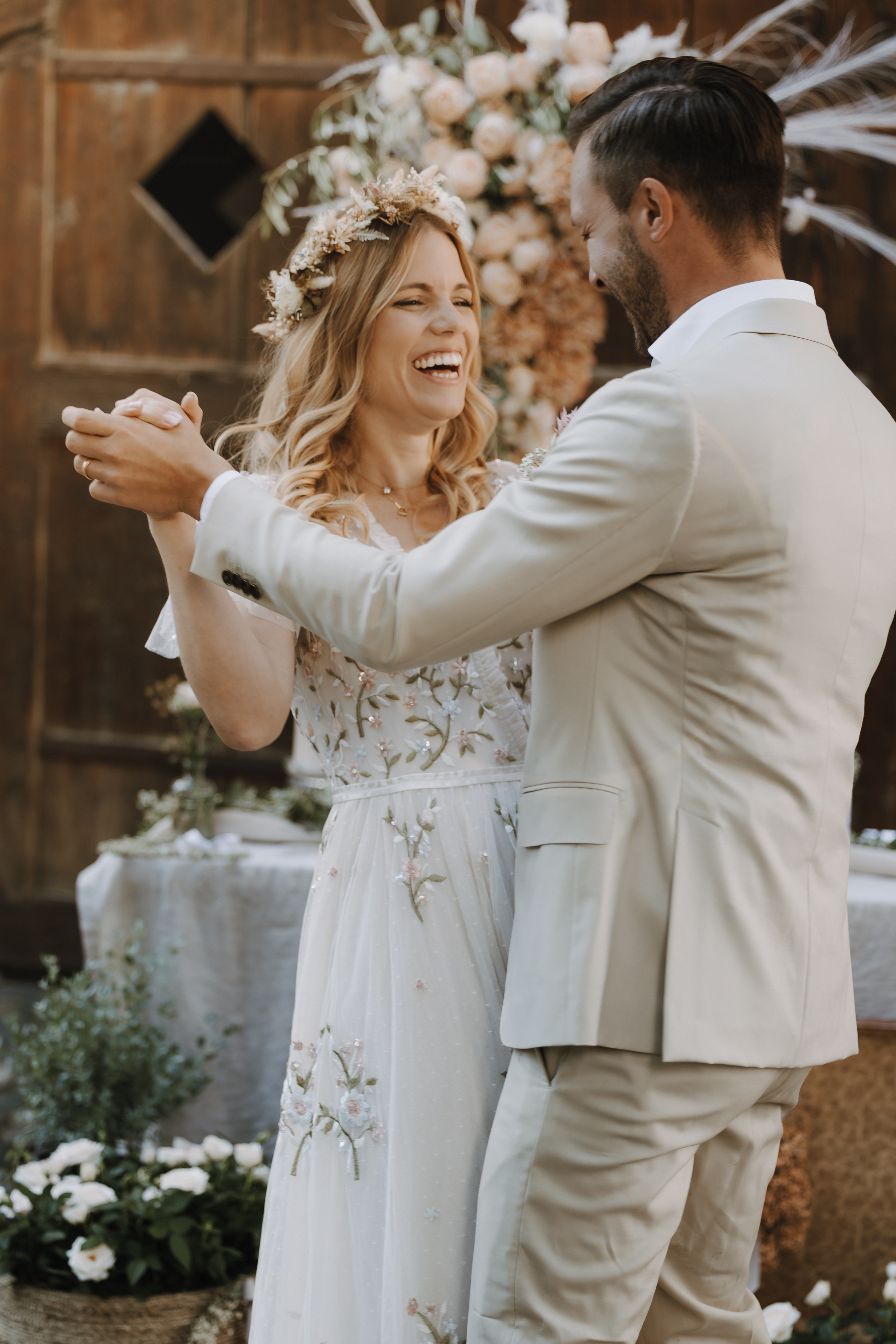 Ein Paar tanzt und lächelt bei einem zauberhaften Fotoshooting bei seiner Hochzeit. Die Frau, geschmückt in einem weißen Kleid mit Blumenmuster und einer Blumenkrone, und der Mann in seinem hellbeigen Anzug sind umgeben von üppigem Grün und Blumendekorationen vor einer bezaubernden Holzkulisse.