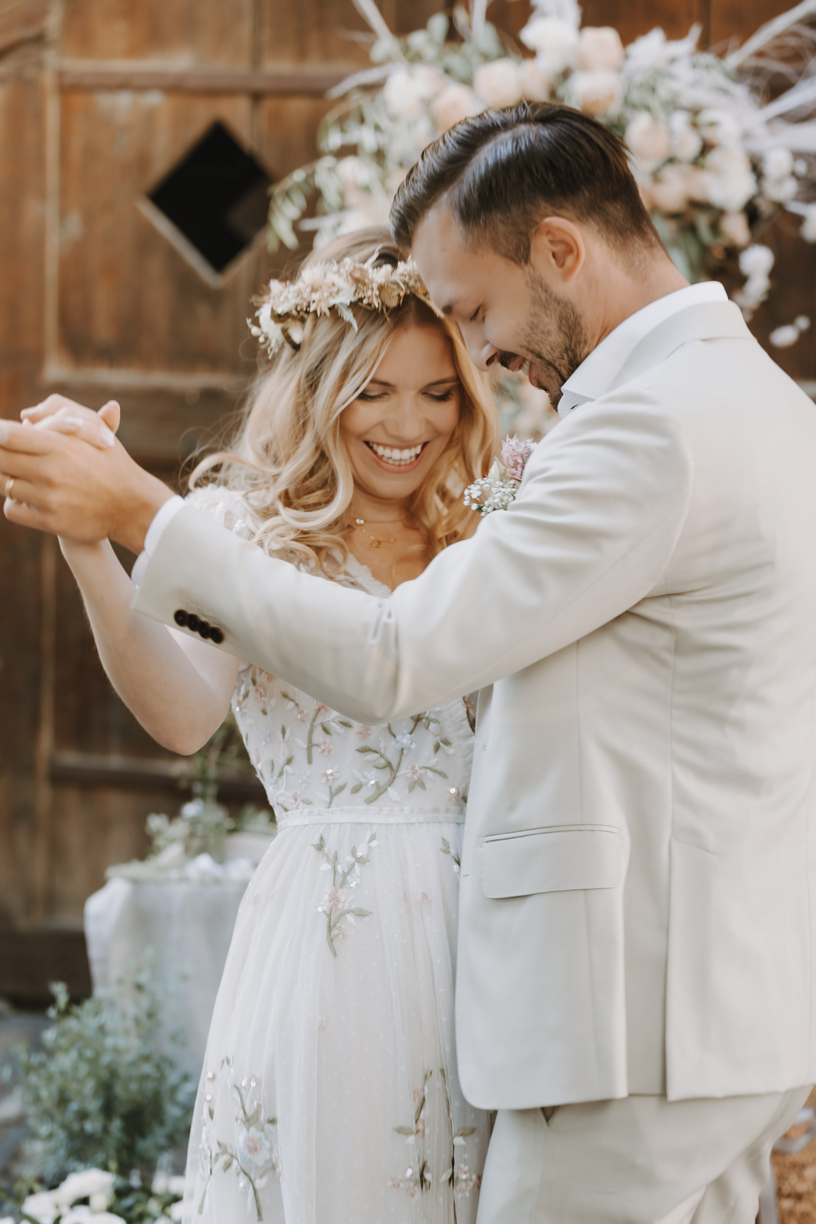 Braut und Bräutigam tanzen fröhlich bei ihrer Hochzeit, wunderschön festgehalten von Hochzeitsfotografin Tasha Ender. Die Braut trägt ein geblümtes Kleid und einen Blumenkranz, während der Bräutigam einen hellen Anzug trägt. Umgeben von elegantem Blumenschmuck teilen sie einen liebevollen Moment.