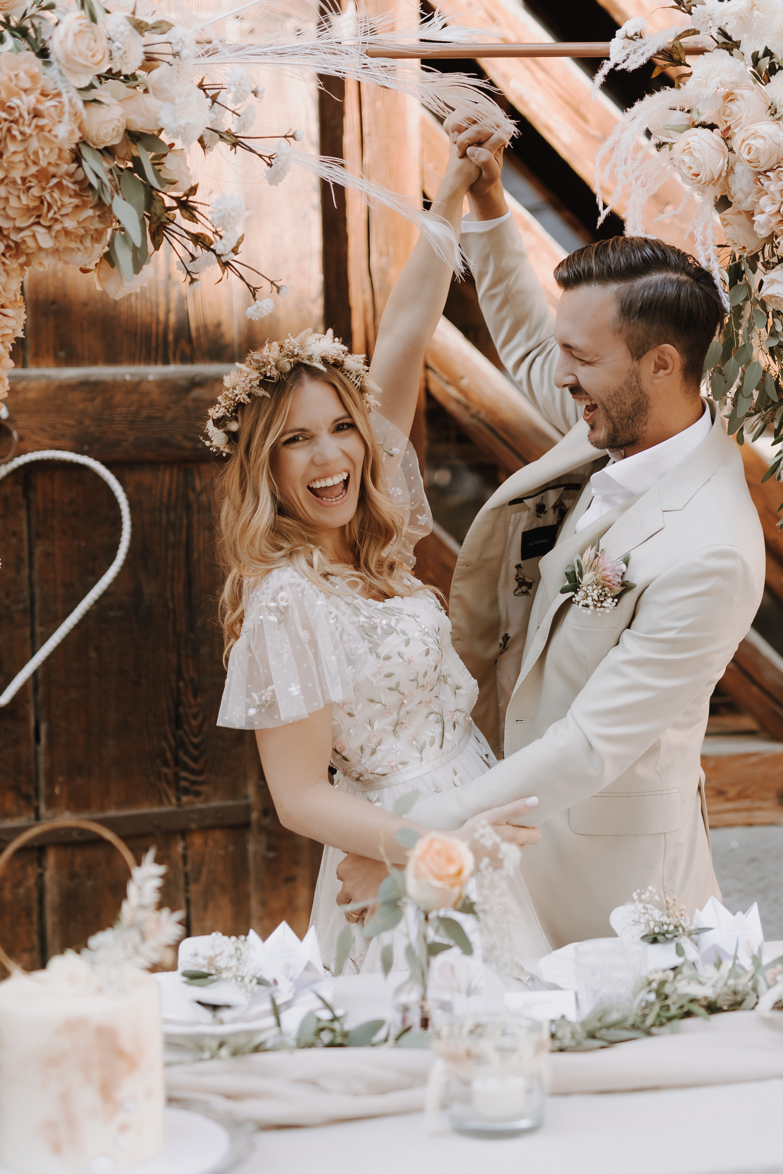 Ein fröhliches Paar in Hochzeitskleidung steht während seiner Hochzeit unter einem Blumenbogen. Sie halten Händchen und lächeln breit. Die Braut ist mit einer Blumenkrone und einem Spitzenkleid geschmückt, während der Bräutigam einen hellen Anzug mit einer Ansteckblume trägt. Der Tisch vor ihnen ist mit Blumen dekoriert.