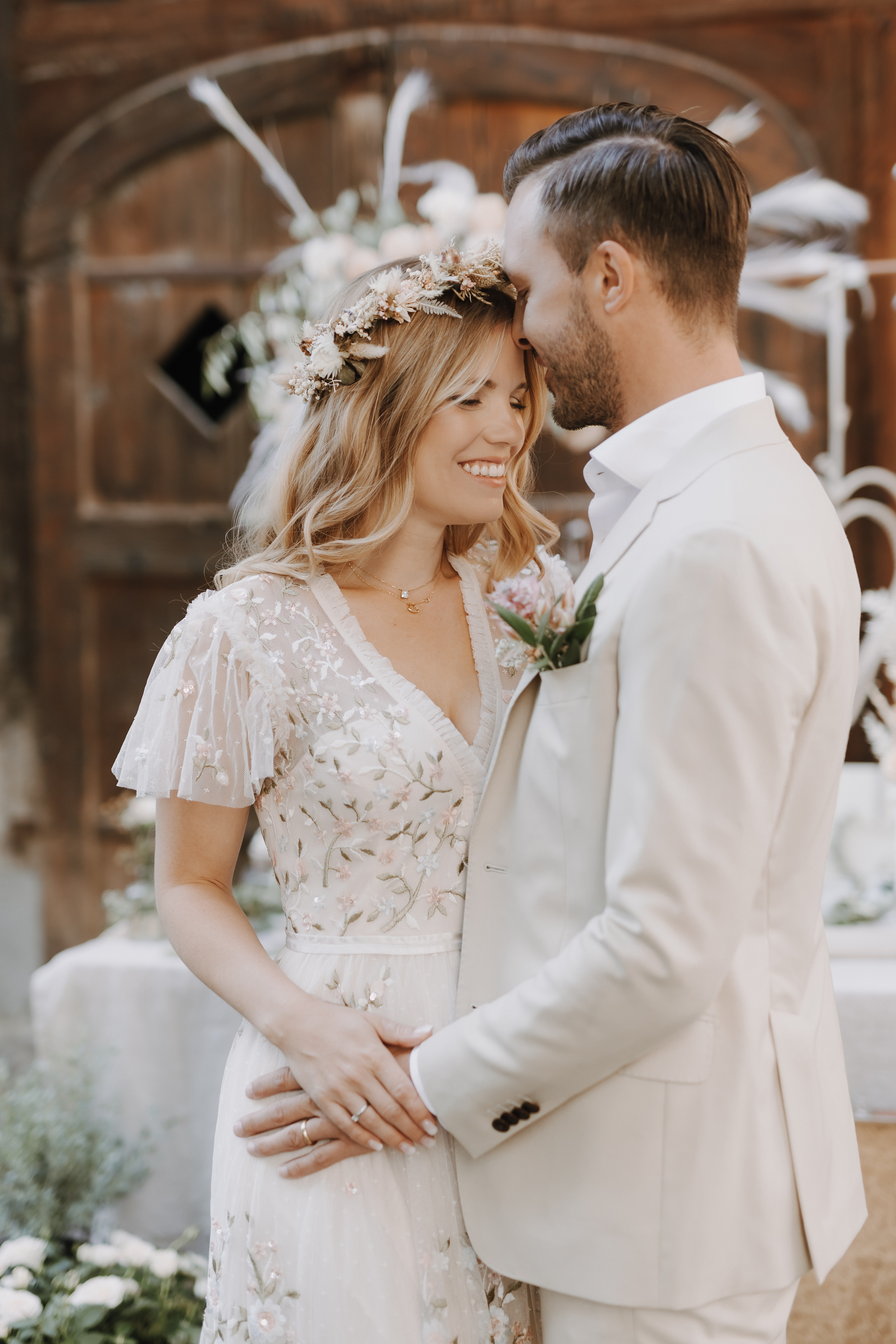 Braut und Bräutigam teilen einen intimen Moment an ihrem Hochzeitstag. Die Braut, die eine Blumenkrone und ein weißes Kleid mit zarten Spitzendetails trägt, lächelt herzlich. Der Bräutigam, der einen hellbeigen Anzug trägt, umarmt sie sanft. Sie stehen vor einer rustikalen Holzkulisse, die von Hochzeitsfotografin Tasha Ender in Baden wunderschön eingefangen wurde.