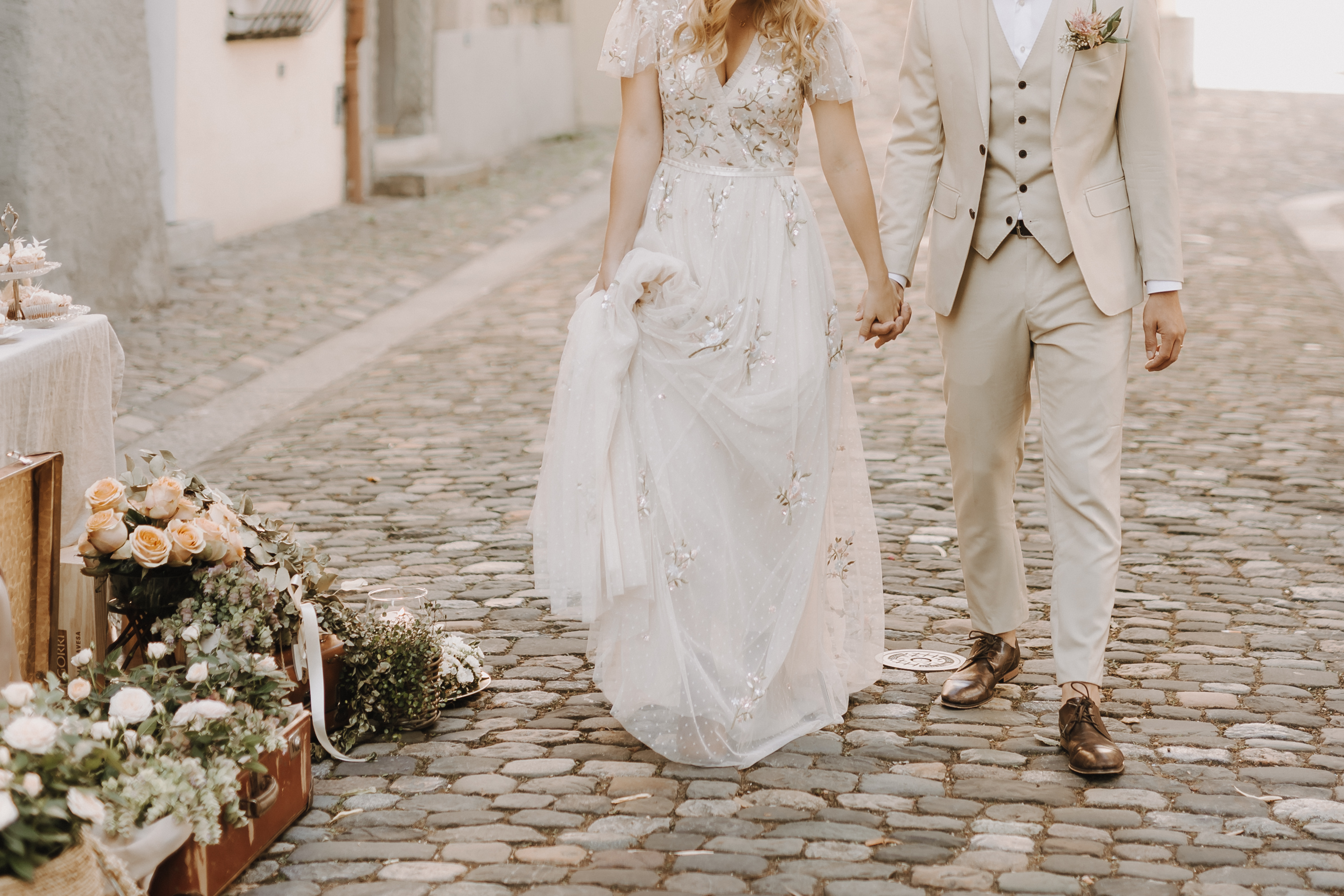 Ein Paar im Hochzeitskleid geht Hand in Hand eine Kopfsteinpflasterstraße entlang. Die Braut, geschmückt mit einem fließenden weißen Kleid mit Blumenverzierungen, und der Bräutigam, gekleidet in einen beigen Anzug, sind von Blumenarrangements und Vintage-Dekor umgeben – eine perfekte Szene für ihre Hochzeitsfotografin, um diesen magischen Moment festzuhalten.