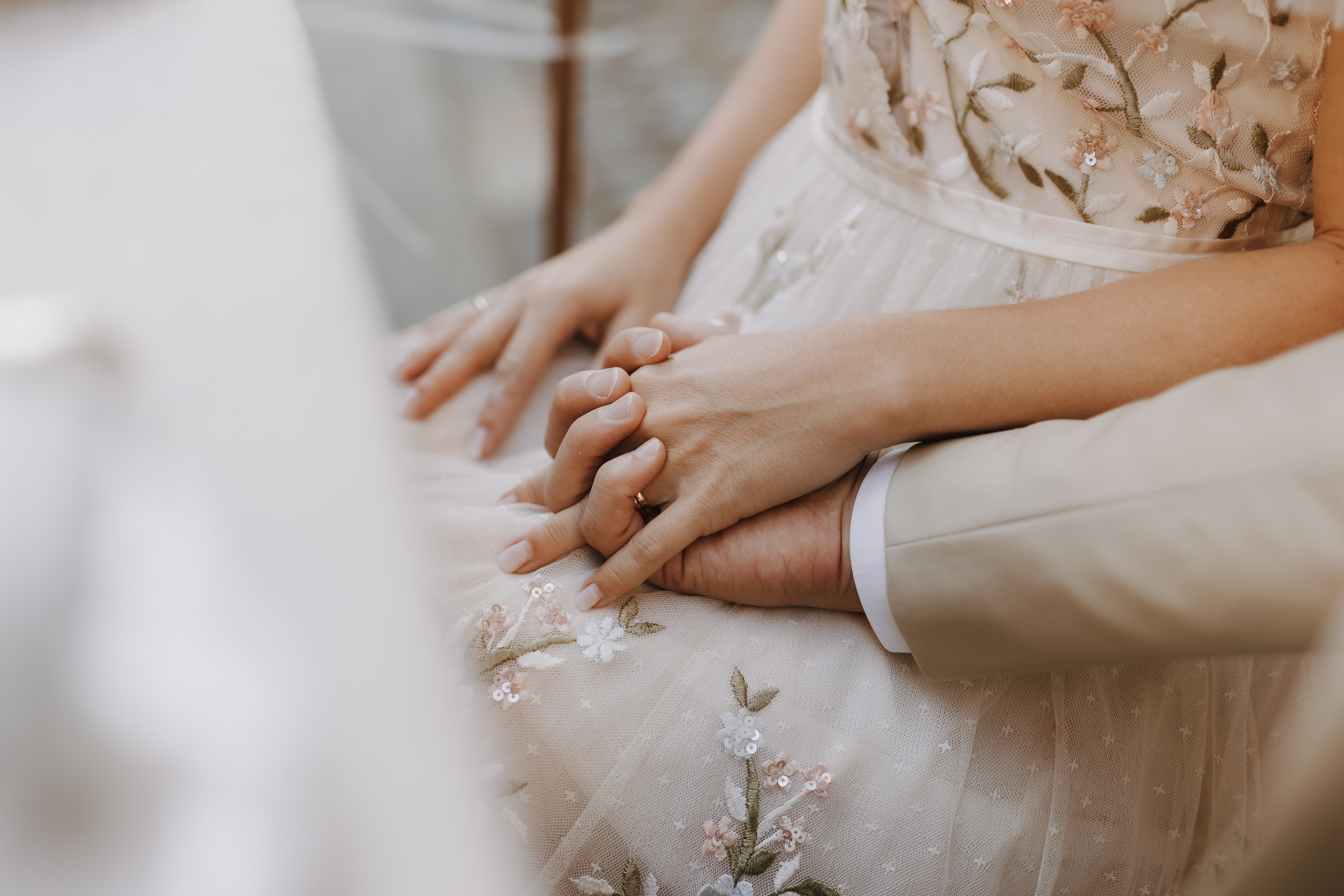 Eine Nahaufnahme eines Paares, das bei seiner Hochzeit Händchen hält. Die Frau, die ein zartes, mit Blumen besticktes Kleid und einen Ring an der linken Hand trägt, steht neben dem Mann, der einen beigen Anzug trägt. Der Fokus liegt auf ihren ineinander verschlungenen Händen, die Intimität und Verbundenheit während ihrer Trauung symbolisieren.