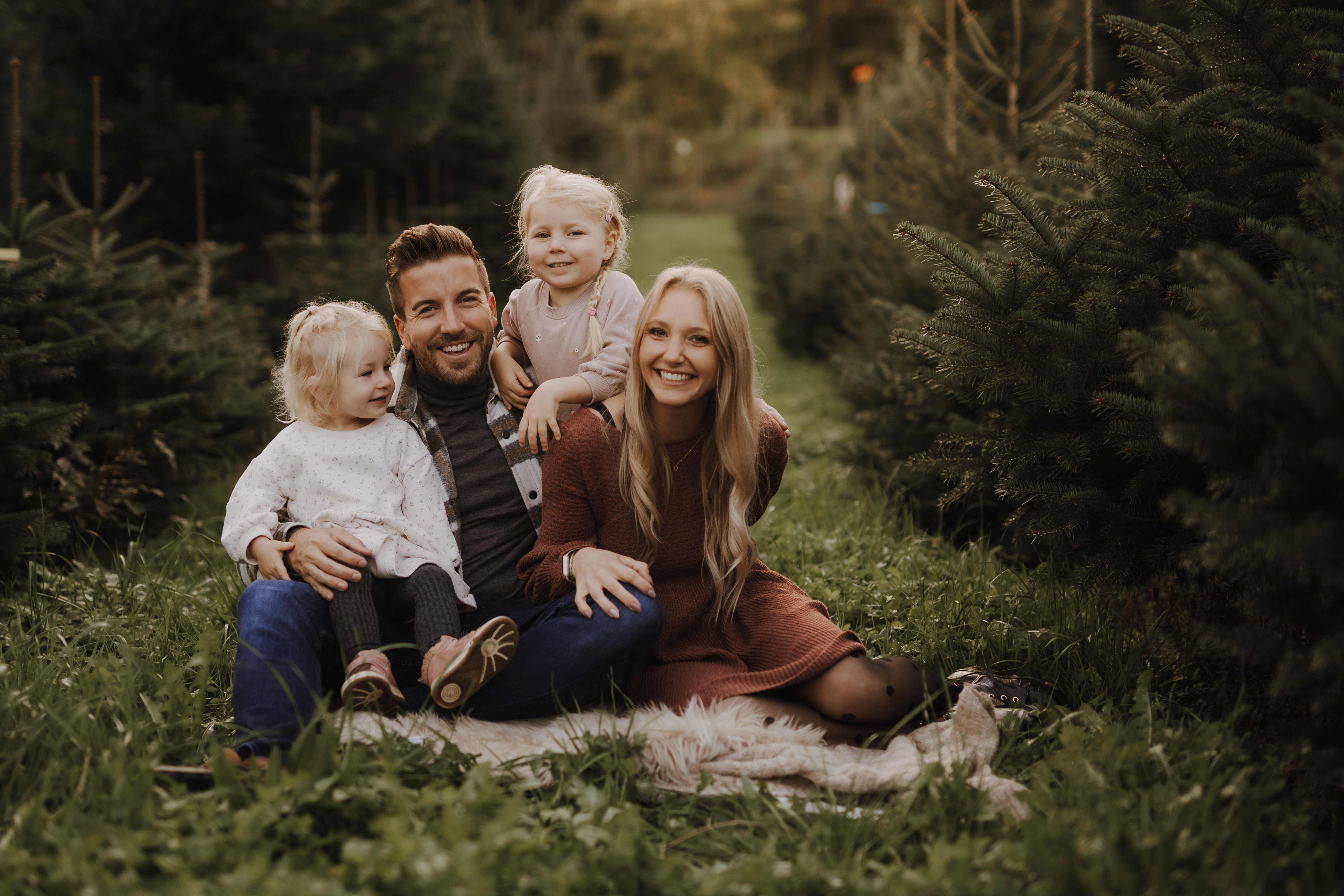 Eine vierköpfige Familie sitzt auf einer Decke in einer von immergrünen Bäumen umgebenen Rasenfläche. Zwei kleine Kinder sitzen auf dem Schoß der lächelnden Eltern, während Vater und Mutter nebeneinander sitzen und einen sonnigen Tag im Freien genießen. Dieser malerische Moment könnte perfekt für ein Familienshooting sein.