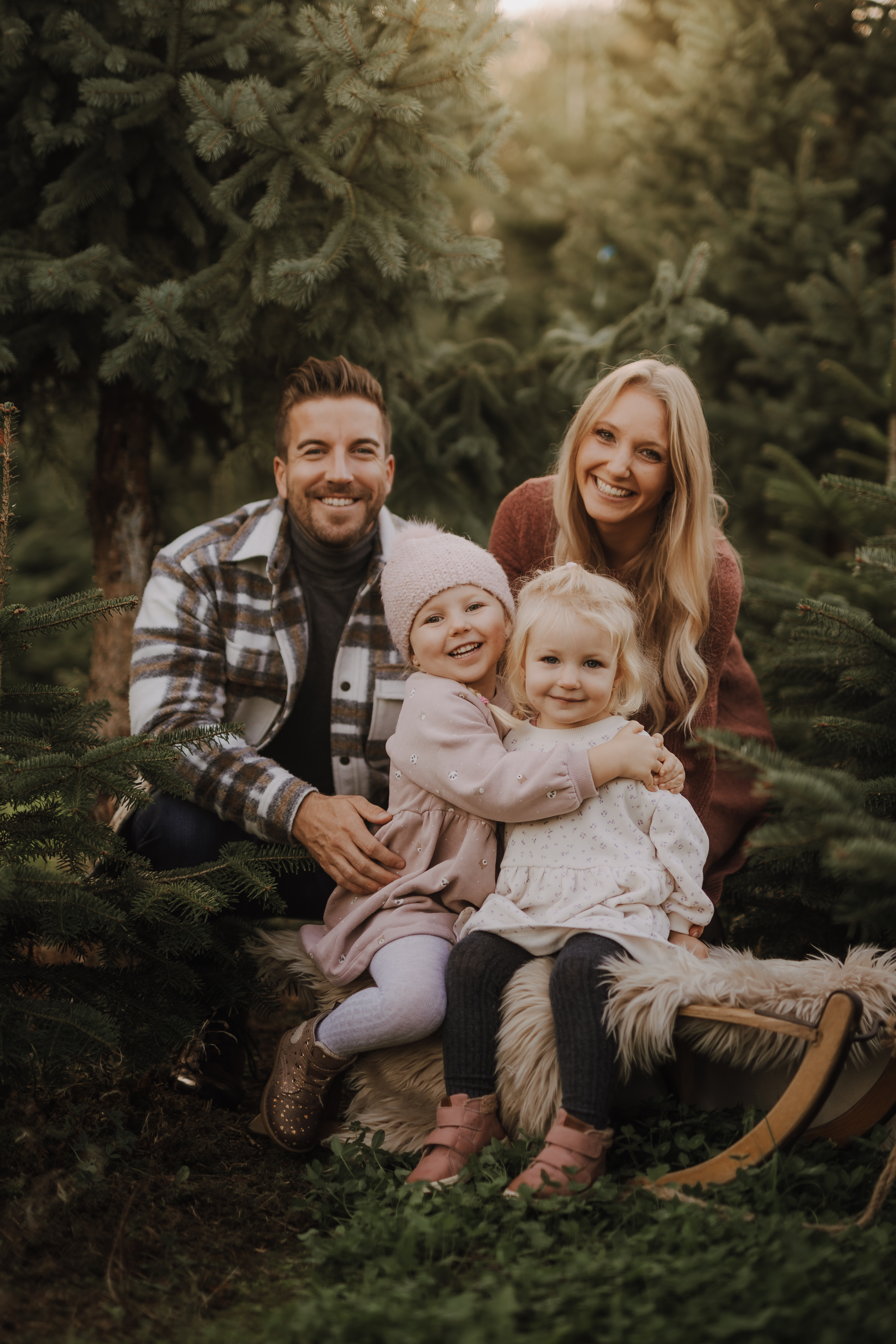 Eine vierköpfige Familie posiert während ihres Weihnachts-Fotoshootings in einem Wald in Baden, umgeben von immergrünen Bäumen. Die lächelnden Eltern knien hinter ihren beiden kleinen Töchtern. Ein Mädchen sitzt auf einer mit Fell bezogenen Bank, während das andere mit einer rosa Strickmütze daneben steht. Alle sind warm angezogen.