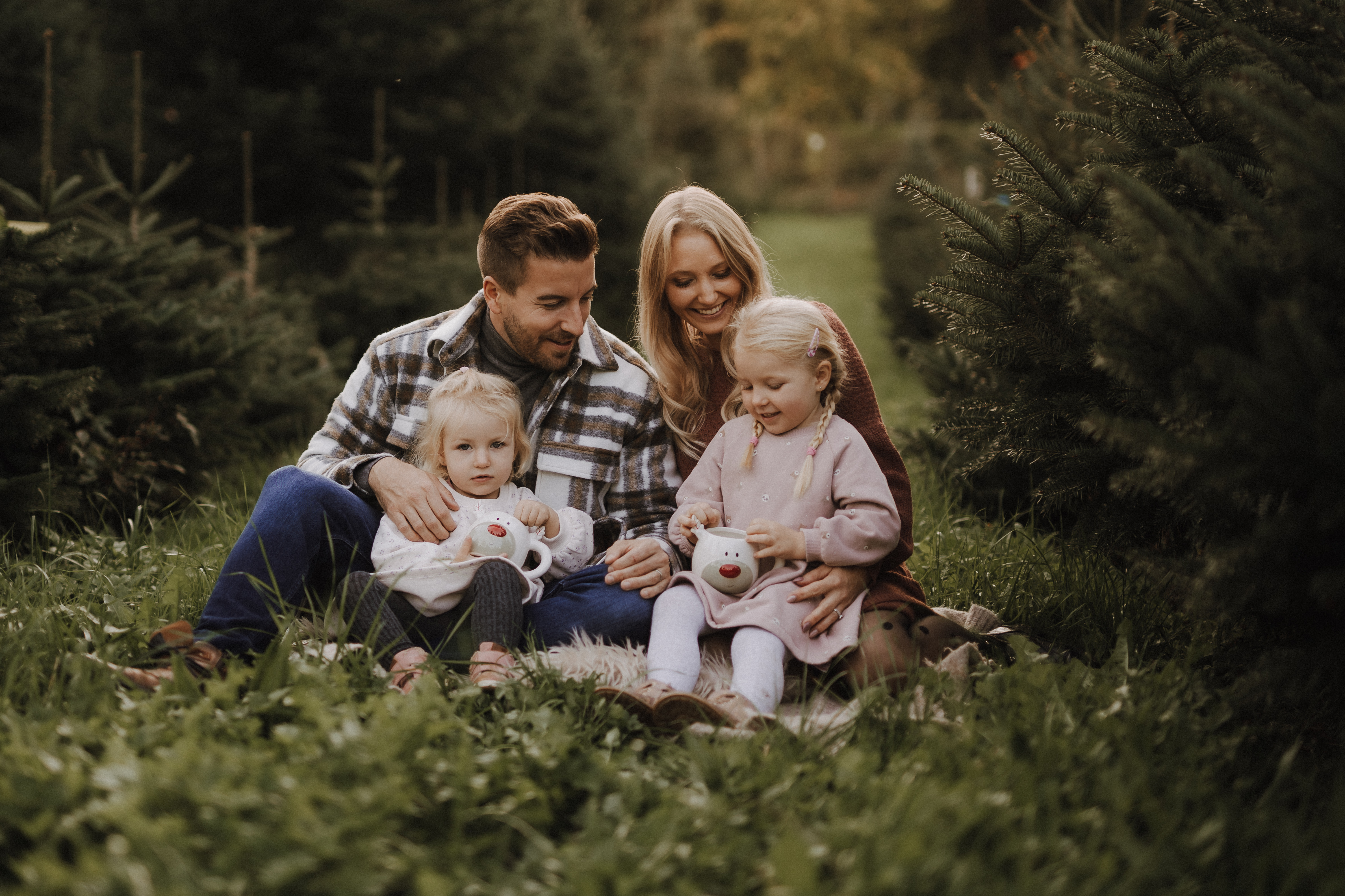 Eine Familie sitzt auf dem Gras in einem Waldstück im Aargau. Ein Mann und eine Frau sitzen eng beieinander und halten jeweils eines ihrer beiden kleinen Kinder. Die Kinder halten Stofftiere. Jeder scheint zu lächeln oder sich auf die Spielzeuge zu konzentrieren. Die Umgebung ist ruhig und grün, perfekt für eine herzerwärmende Fotoshooting-Aktion.