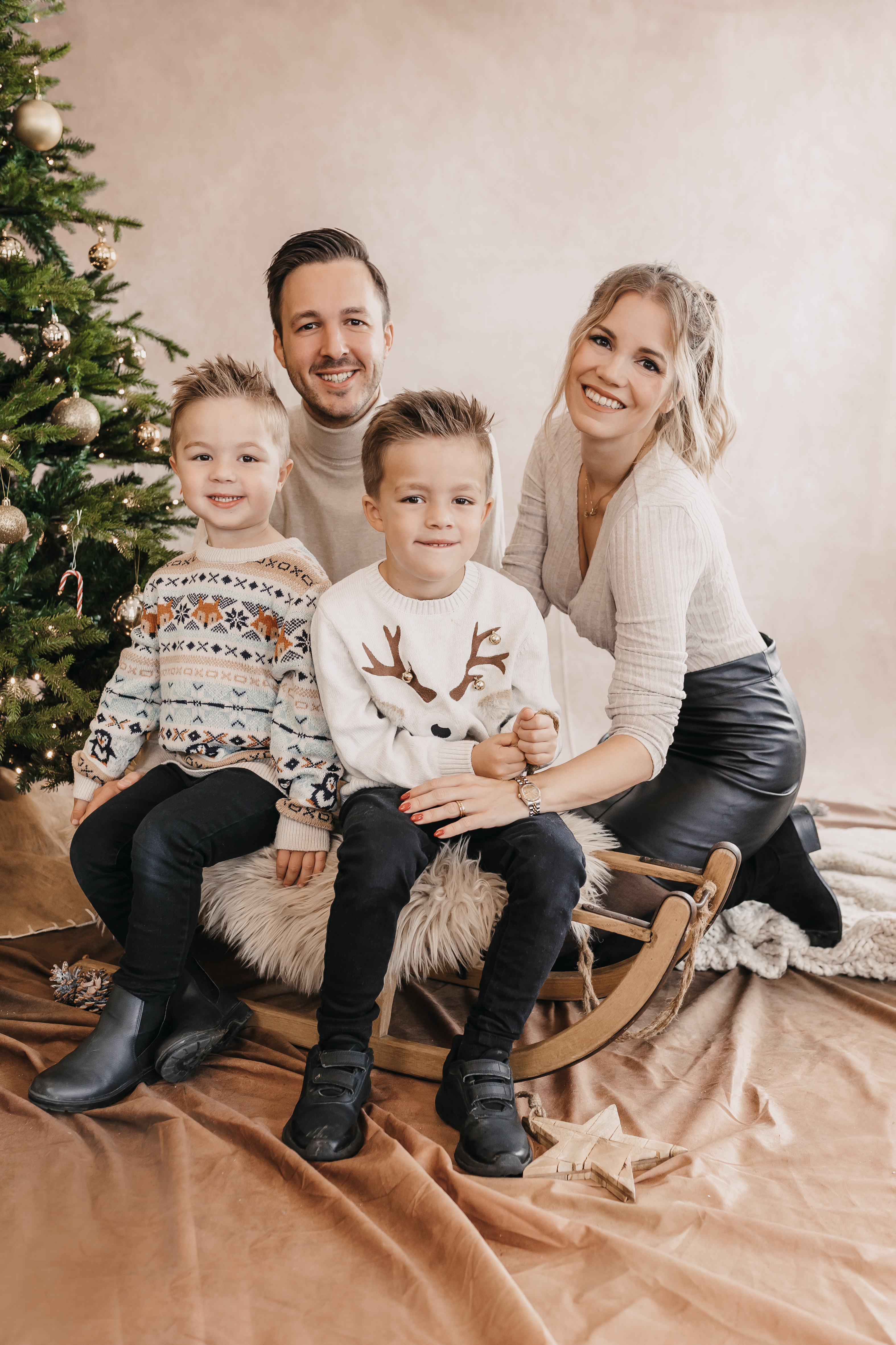 Eine glückliche vierköpfige Familie posiert während ihres Familienshootings neben einem geschmückten Weihnachtsbaum. Mutter und Vater knien hinter ihren beiden kleinen Söhnen, die auf einem Holzschlitten mit einer Felldecke sitzen. Alle sind in kuschelige Pullover gekleidet und lächeln herzlich in die Kamera. Die Kulisse hat eine festliche und warme Atmosphäre.