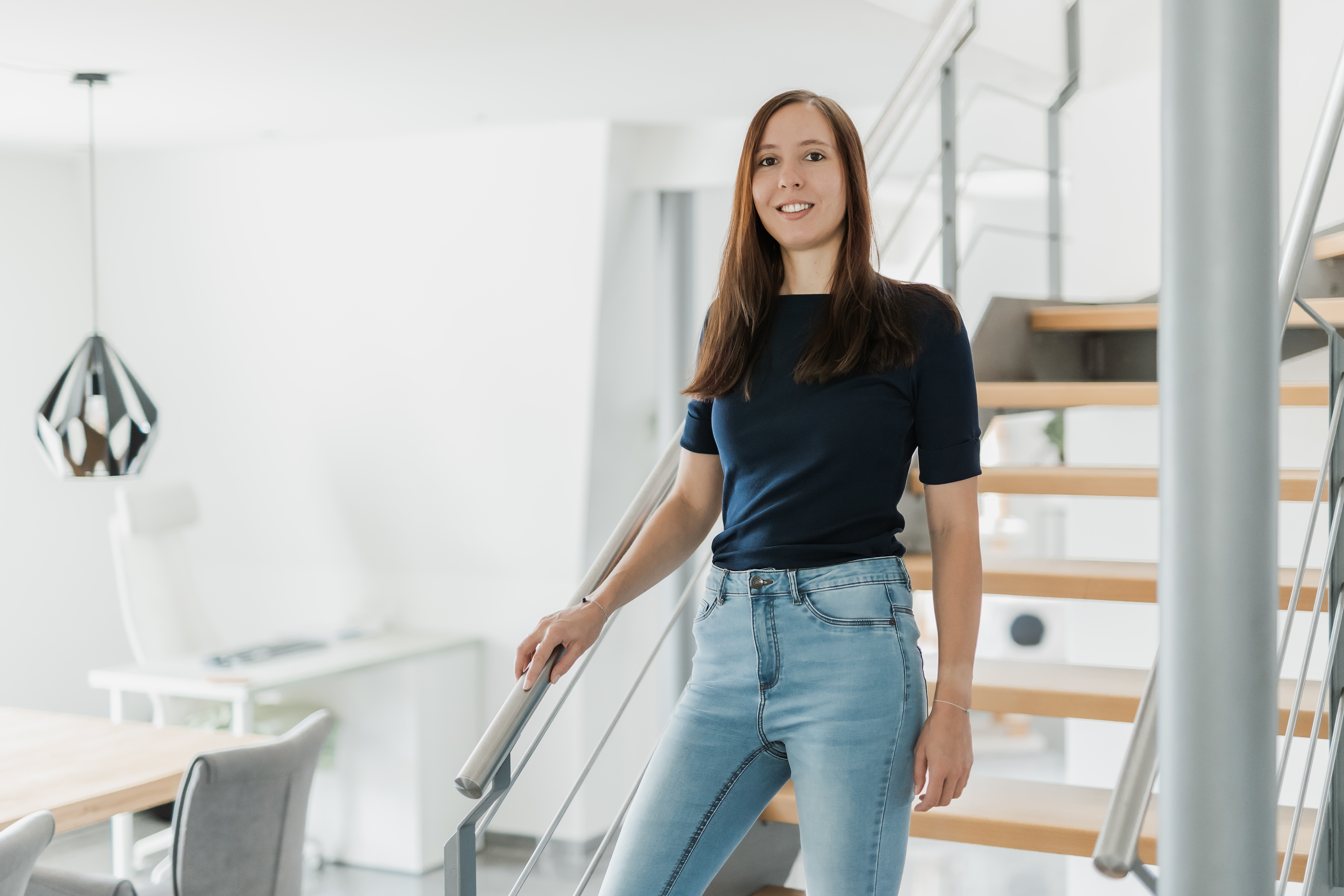 Eine Frau mit langen braunen Haaren, einem marineblauen Oberteil und hellblauen Jeans trägt, steht selbstbewusst auf einer modernen Holztreppe in einem hellen, minimalistischen Wohninterieur und strahlt die gelassene Eleganz eines professionellen Porträts aus, das sich ideal für jeden Lebenslauf eignet.