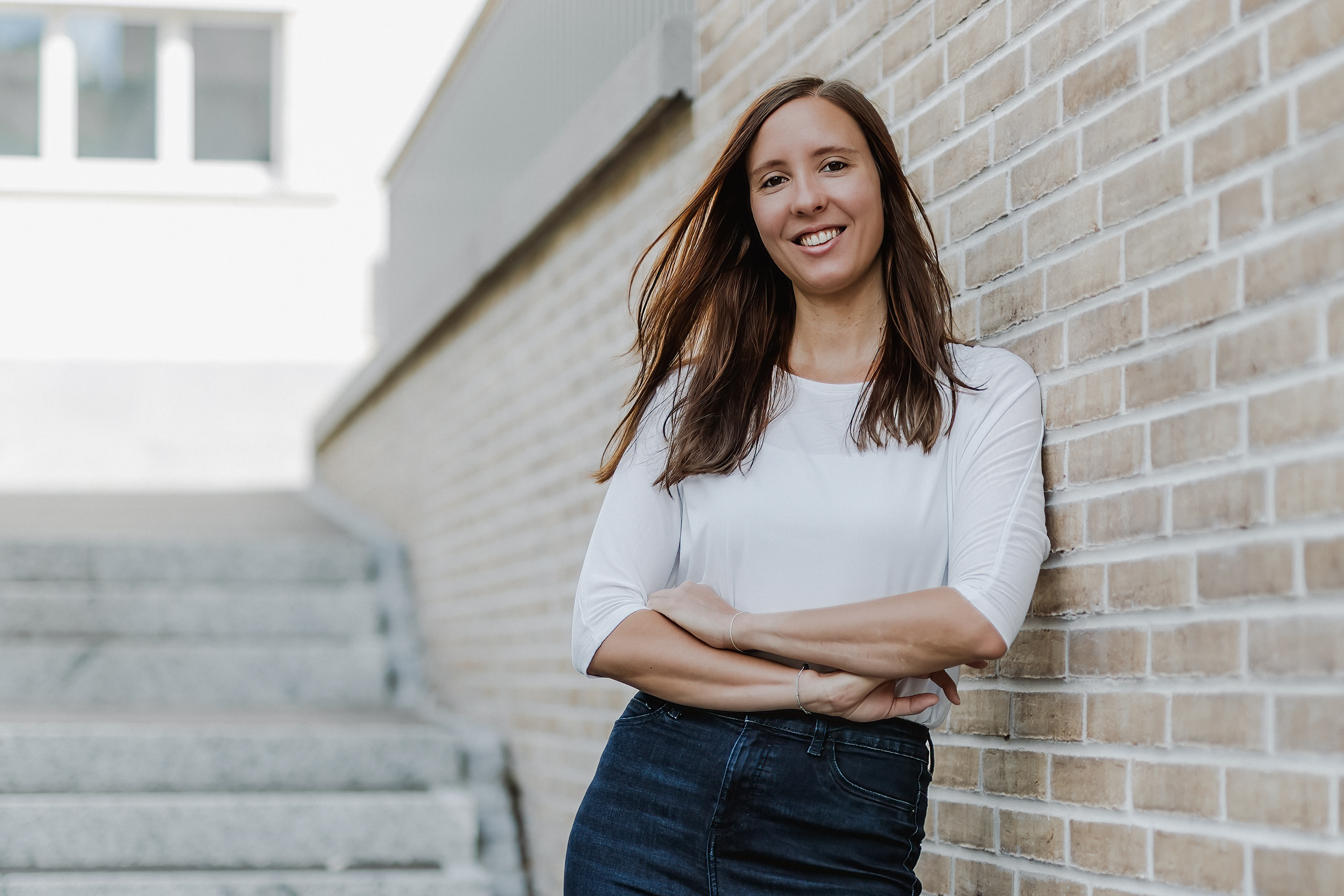 Eine Frau mit langen braunen Haaren lehnt lächelnd an einer Backsteinmauer. In ihrem weißen Hemd und dunklen Jeans strahlt sie Selbstbewusstsein aus – und verkörpert damit das perfekte Porträt für ihren Lebenslauf. Im Hintergrund führen Stufen nach oben, während das Sonnenlicht sanfte Schatten wirft.