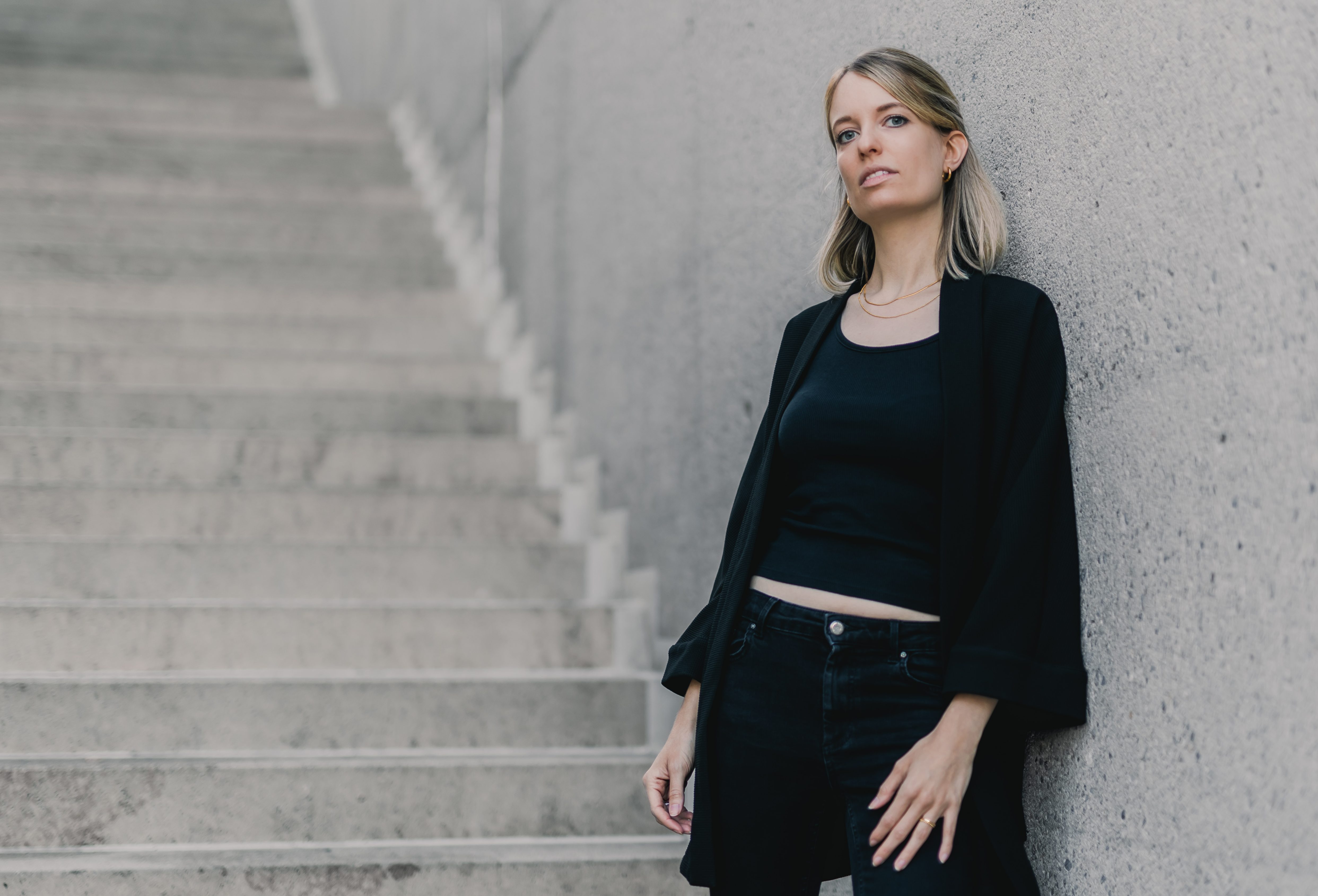 Eine Person mit langen Haaren steht in schwarzem Outfit an einer Betonwand neben einer Treppe. Die Kulisse ist urban und die Stimmung besinnlich, perfekt für ein professionelles Portrait. Die Treppe führt nach oben aus dem Bild heraus.