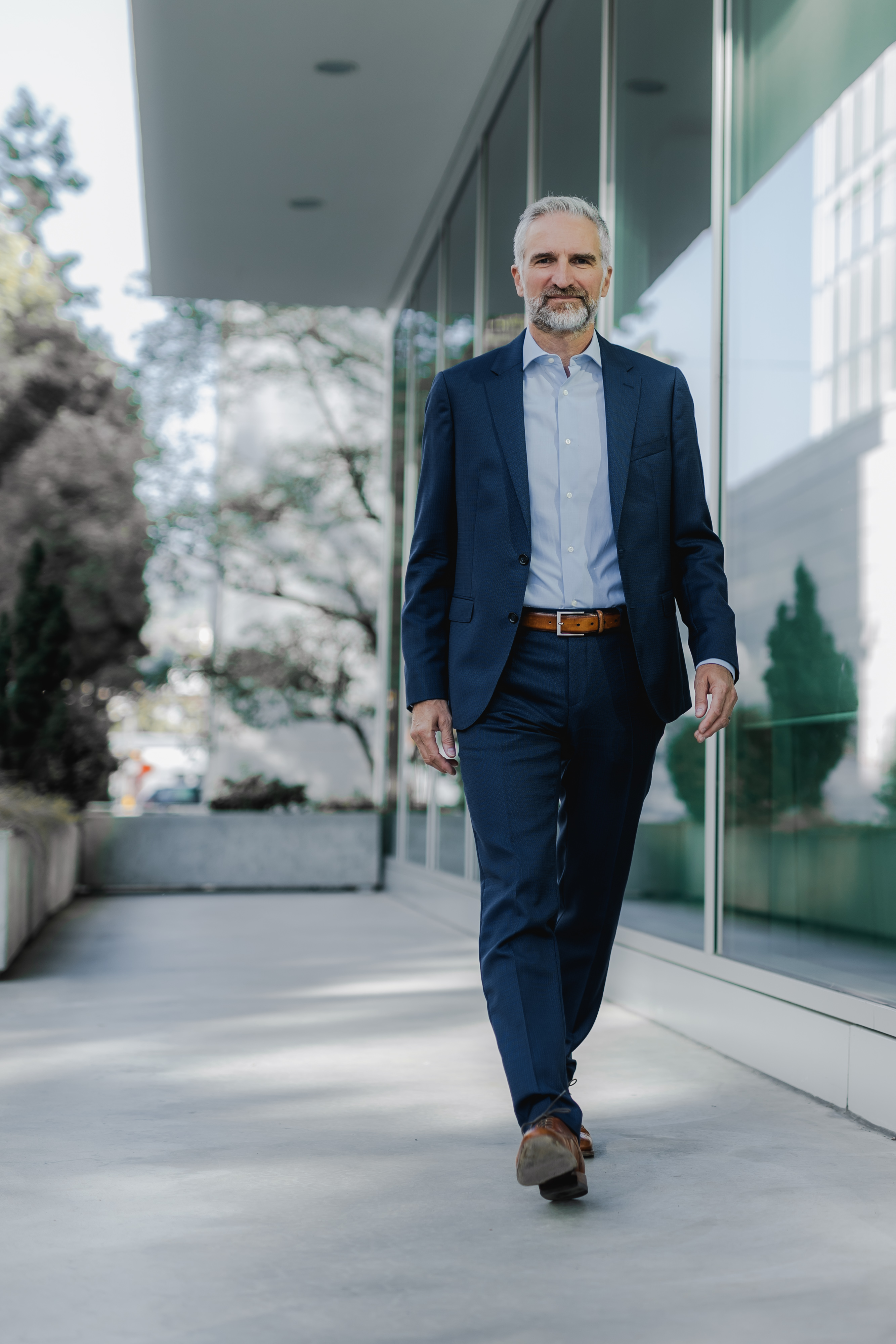Ein Mann mit grauem Haar und Bart geht an einem modernen Gebäude mit Glasfenstern entlang und fängt die Essenz eines professionellen Bewerbungsfotos ein. In einem marineblauen Anzug, einem hellblauen Hemd und braunen Schuhen strahlt er inmitten der Bäume und des Grüns im Hintergrund Selbstvertrauen aus.