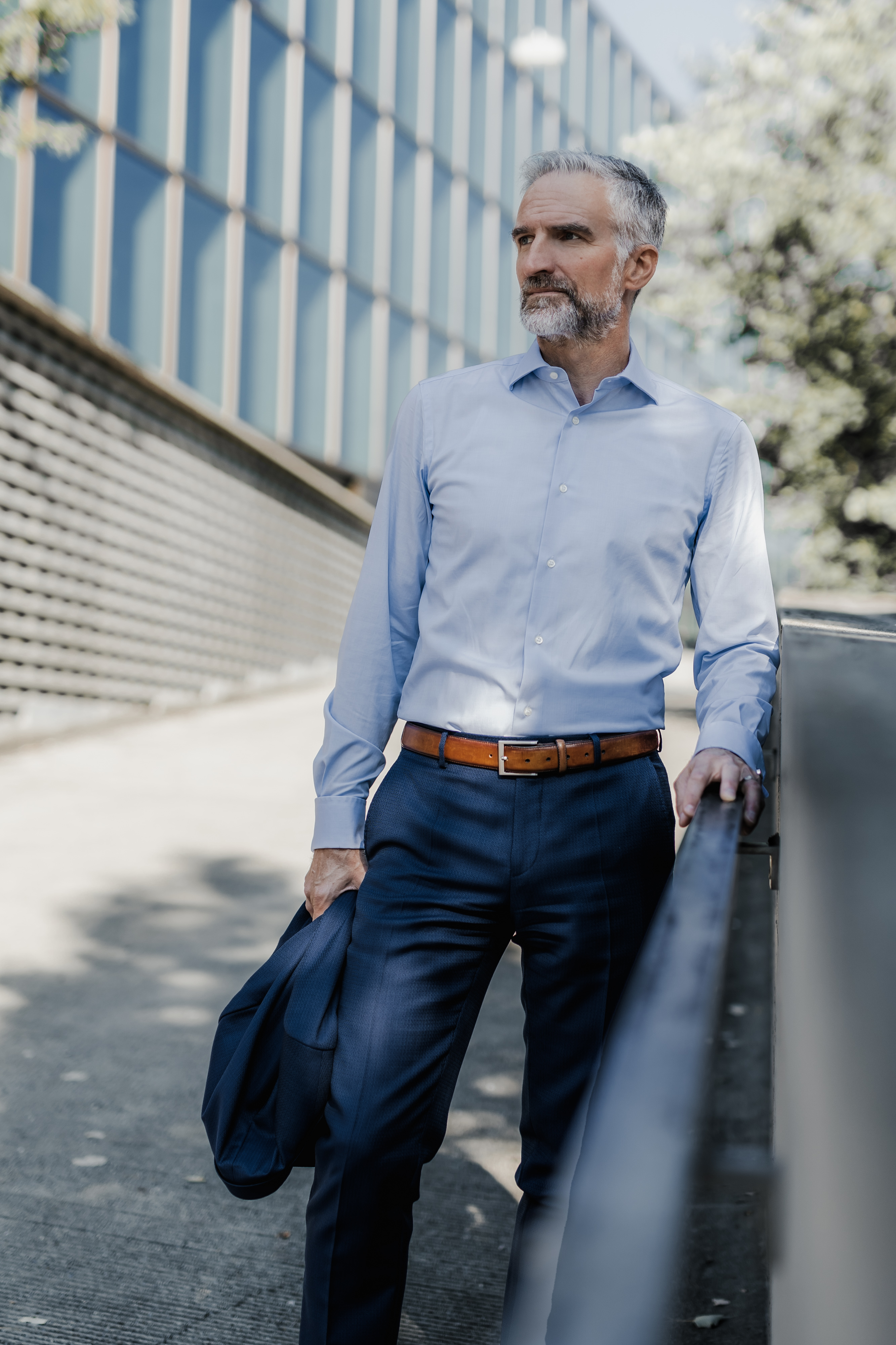 Ein reifer Mann mit grauem Haar und Bart steht auf einem Gehweg und projiziert das perfekte Lebenslauf-Porträt. Er hält eine Jacke in der Hand und trägt ein hellblaues Hemd mit dunkelblauen Hosen. Der Hintergrund – ein modernes Gebäude mit Glasscheiben und Bäumen – ergänzt dieses professionelle Ambiente.