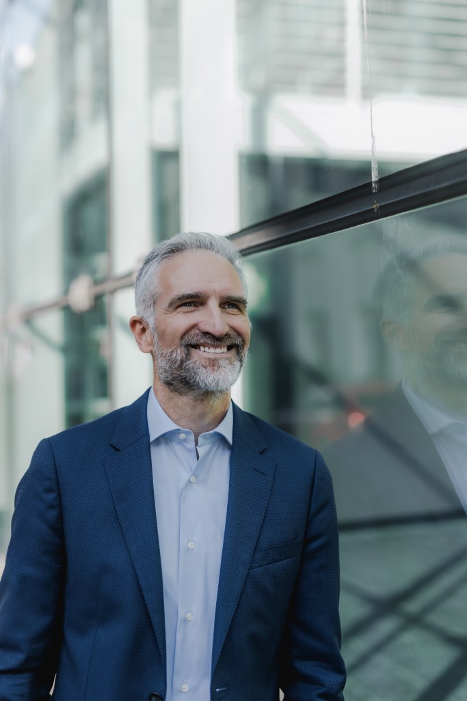 Ein Mann mit grauem Haar und Bart, gekleidet in einen blauen Anzug und ein weißes Hemd, lächelt im Freien für ein Geschäftsporträt. Er steht neben einer Glaswand, die sein Bild vor dem hellen Hintergrund reflektiert, auf dem ein verschwommenes Gebäude zu sehen ist.