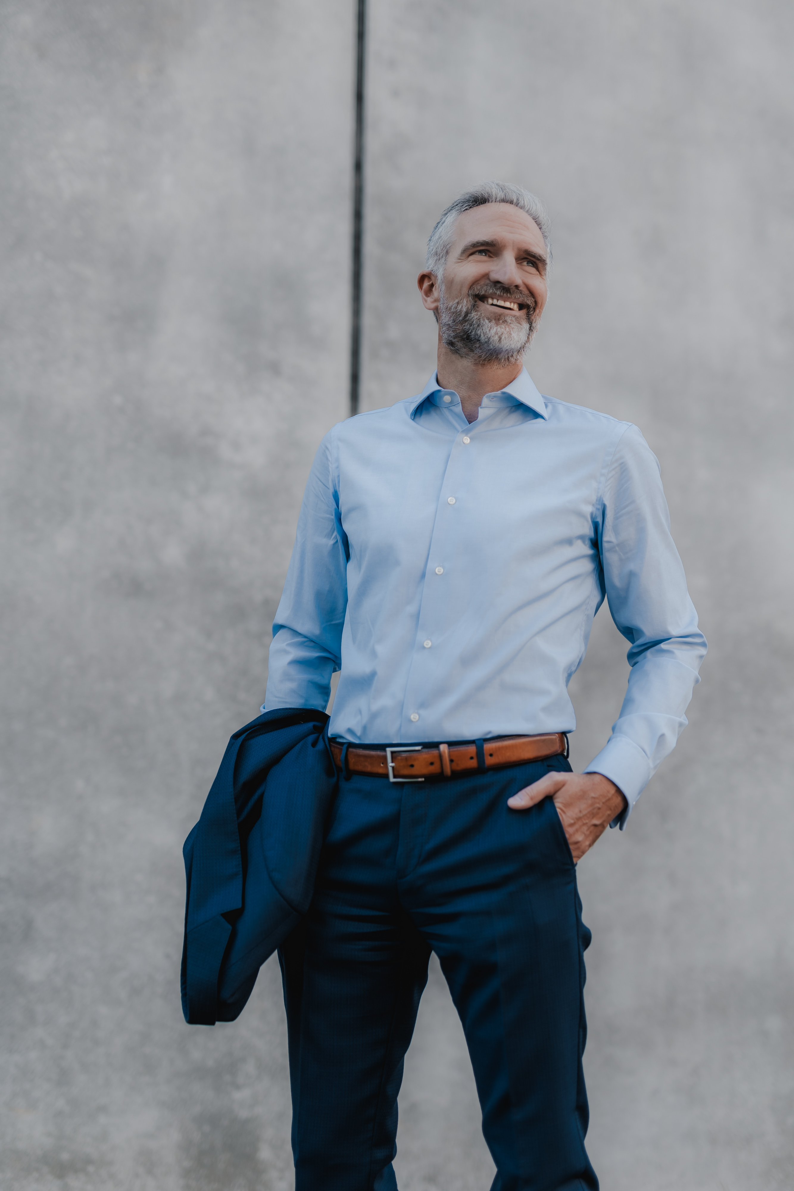 Ein lächelnder Mann mit grauem Haar und Bart posiert für sein Lebenslauf-Porträt vor einer Betonwand. Er trägt ein hellblaues Hemd und dunkle Hosen, ergänzt durch einen braunen Gürtel. Sein Jackett hängt über seinem rechten Arm, während seine linke Hand lässig in seiner Tasche ruht.