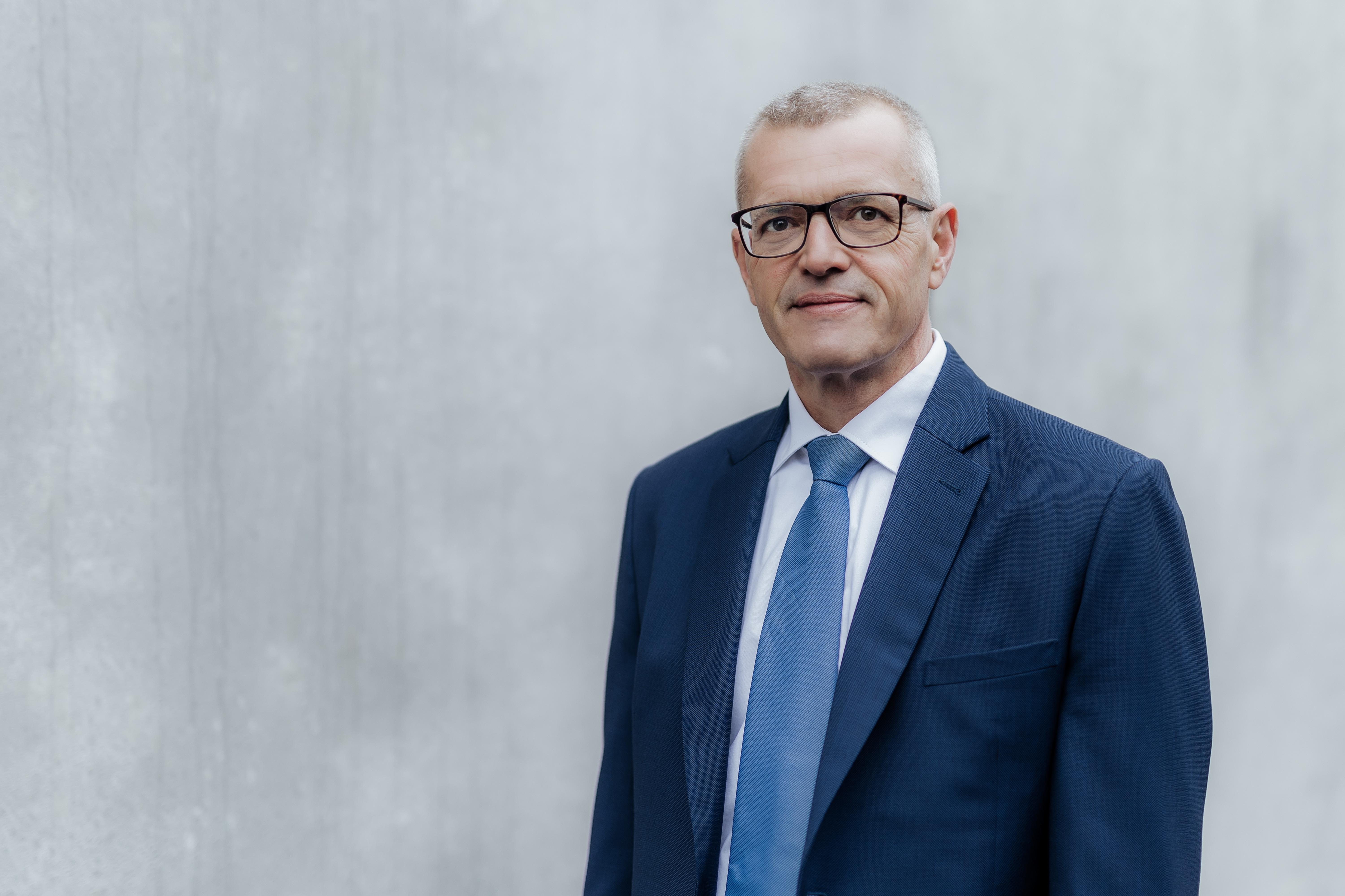 Ein Mann mit Brille und blauem Anzug mit passender Krawatte steht vor einem schlichten, hellen Hintergrund. Dieses professionelle Porträt zeigt ihn mit ruhigem Gesichtsausdruck direkt in die Kamera – ideal für Bewerbungen im Lebenslauf.