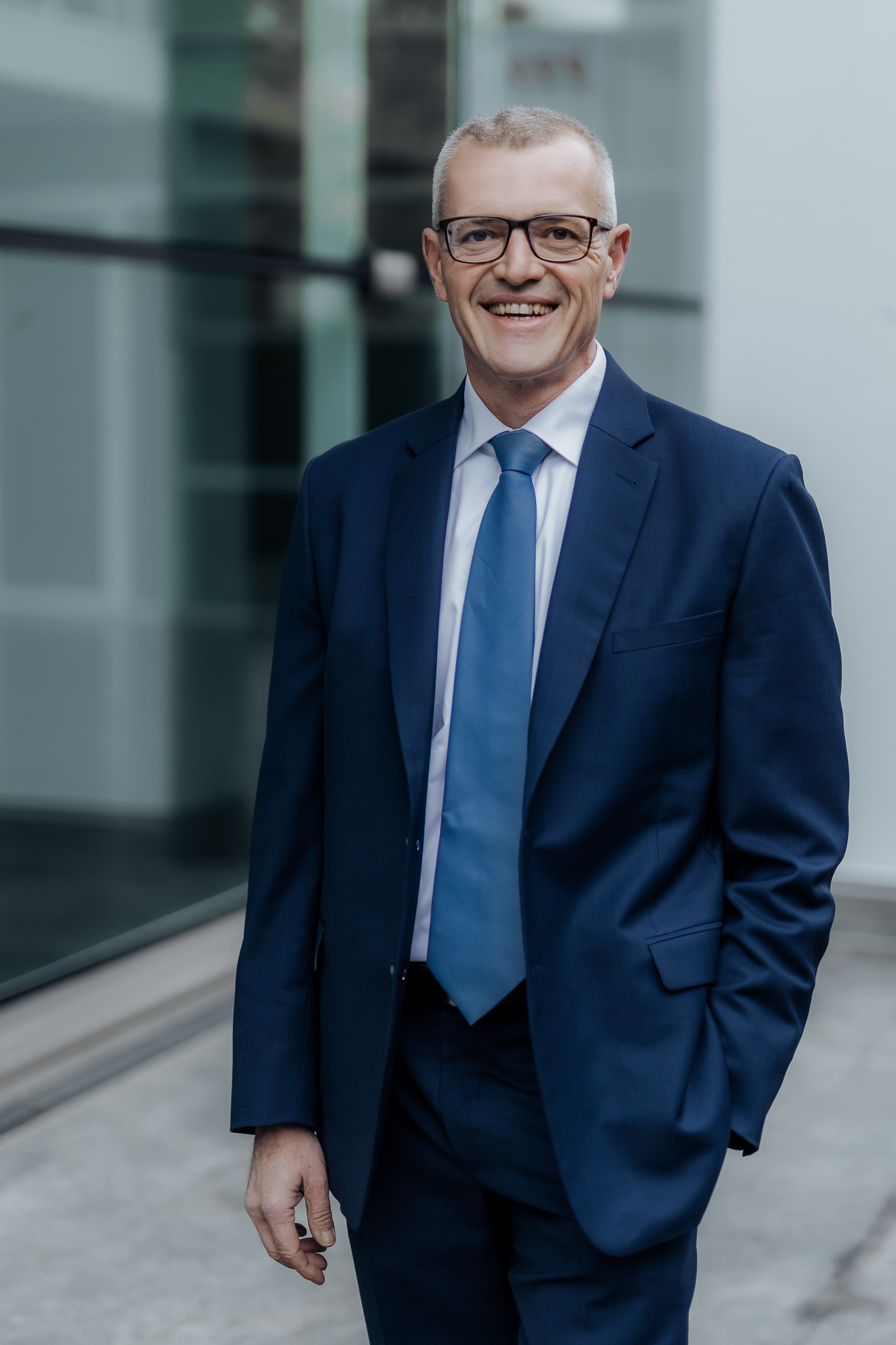 Ein lächelnder Mann in einem blauen Anzug und einer hellblauen Krawatte steht im Freien vor einem modernen Glasgebäude und fängt die Essenz eines professionellen Porträts ein, das sich ideal für einen Lebenslauf oder ein Businessporträt eignet.