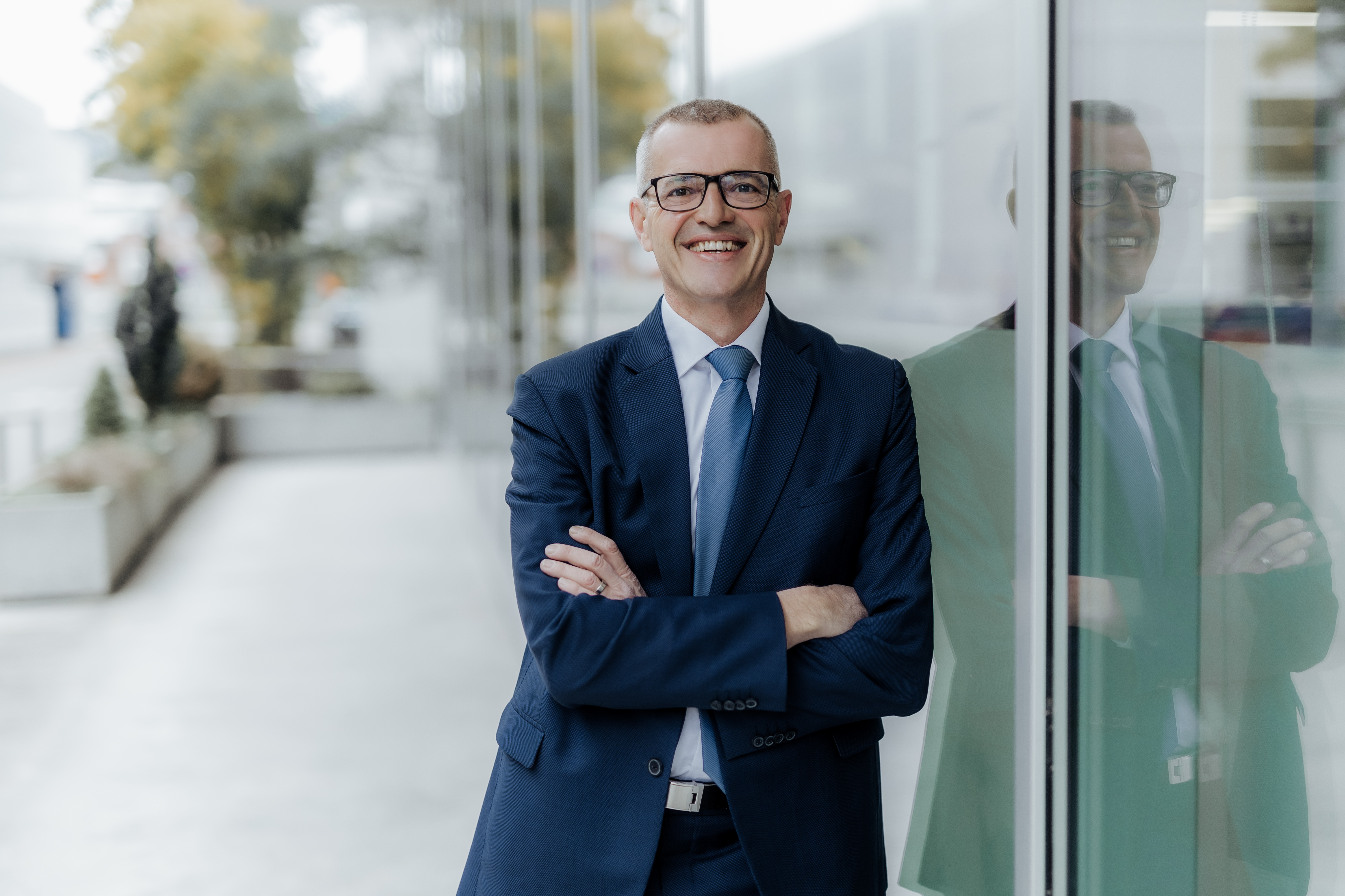 Ein Mann in blauem Anzug und Brille steht selbstbewusst mit verschränkten Armen lächelnd vor einer Glaswand. Die Spiegelung hebt dieses CV-Porträt vor einer verschwommenen städtischen Außenkulisse im Hintergrund hervor.