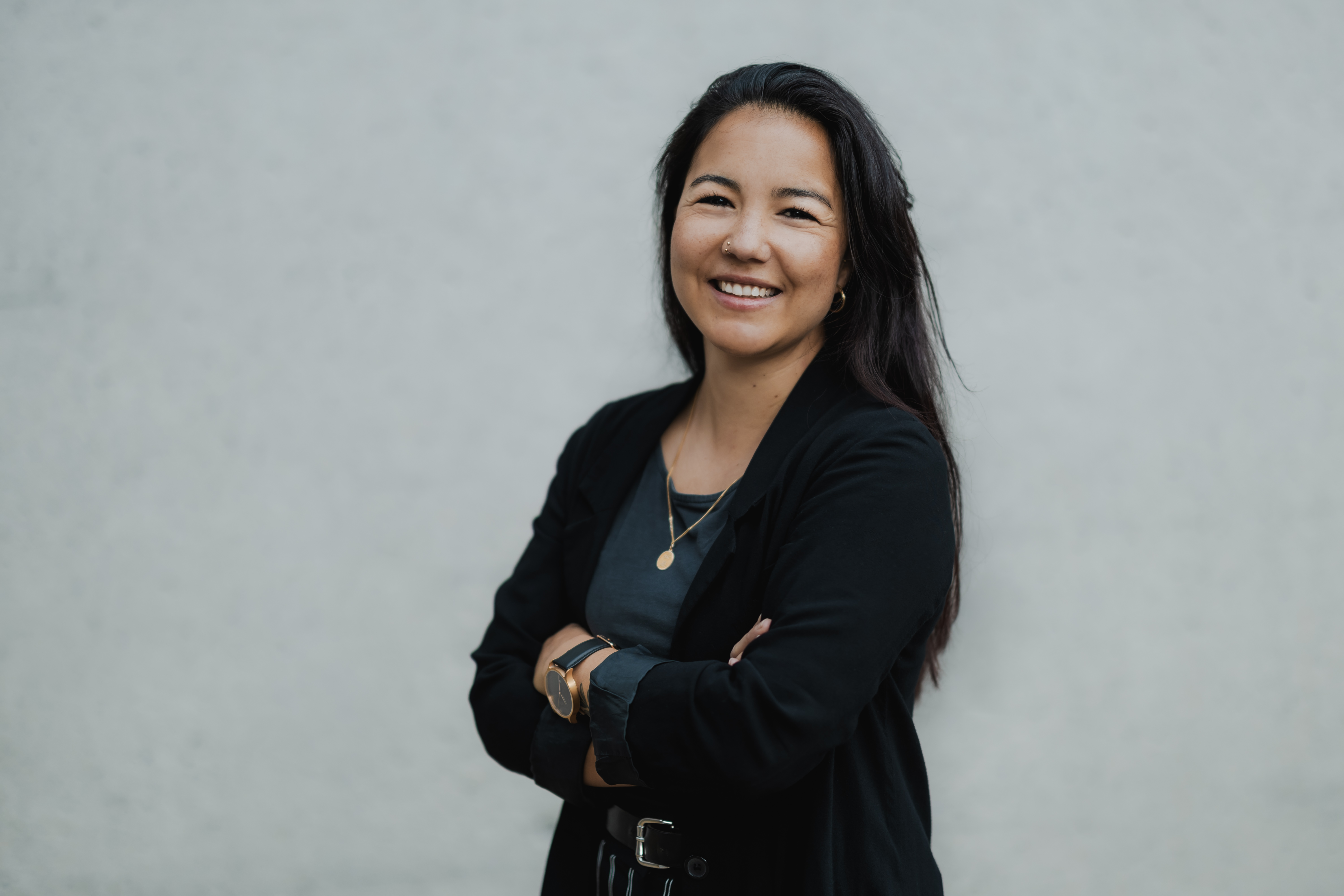 Eine Frau mit langen dunklen Haaren lächelt selbstbewusst in die Kamera. Sie steht vor einem schlichten grauen Hintergrund. Sie trägt einen schwarzen Blazer über einem dunklen Oberteil und hat die Arme in einer entspannten Pose verschränkt – ein ideales Businessportrait für ihren Lebenslauf.