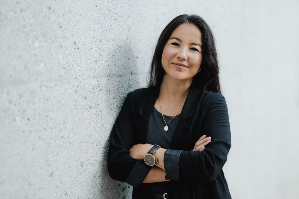 Eine Frau mit langen dunklen Haaren steht selbstbewusst mit verschränkten Armen an einer hellen Wand. Sie trägt einen schwarzen Blazer und ein dunkles Top und ihr subtiles Lächeln zeugt von der Kraft eines professionellen Businessportraits, das jeden Lebenslauf aufwertet. Der sanft unscharfe Hintergrund verleiht ihm eine elegante Note.