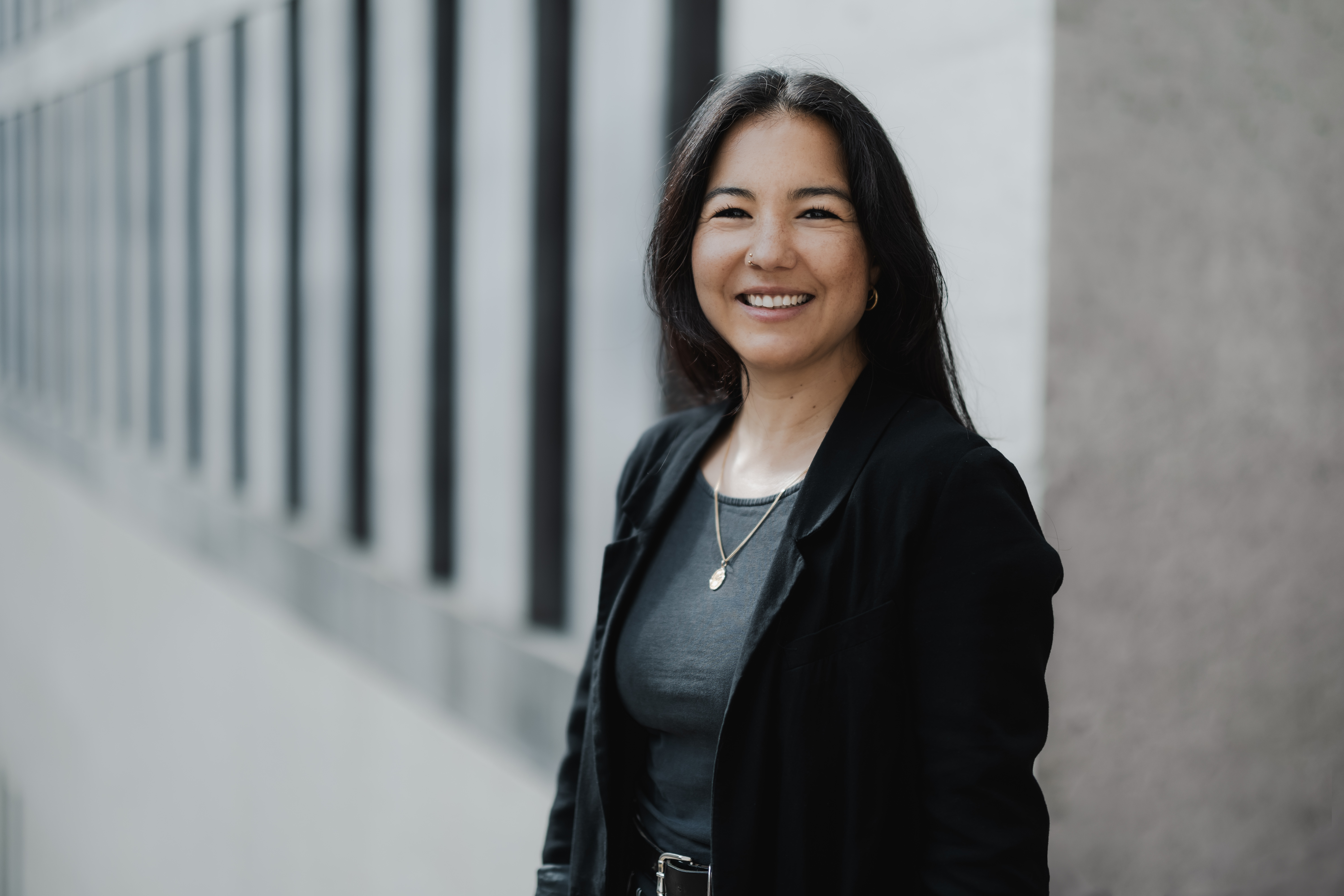 Eine Frau mit langen dunklen Haaren lächelt für ein perfektes CV-Portrait und steht neben einem Gebäude mit großen Fenstern. Sie trägt einen schwarzen Blazer und ein dunkles Oberteil, dazu Halsketten. Der Hintergrund fängt eine städtische Umgebung ein.
