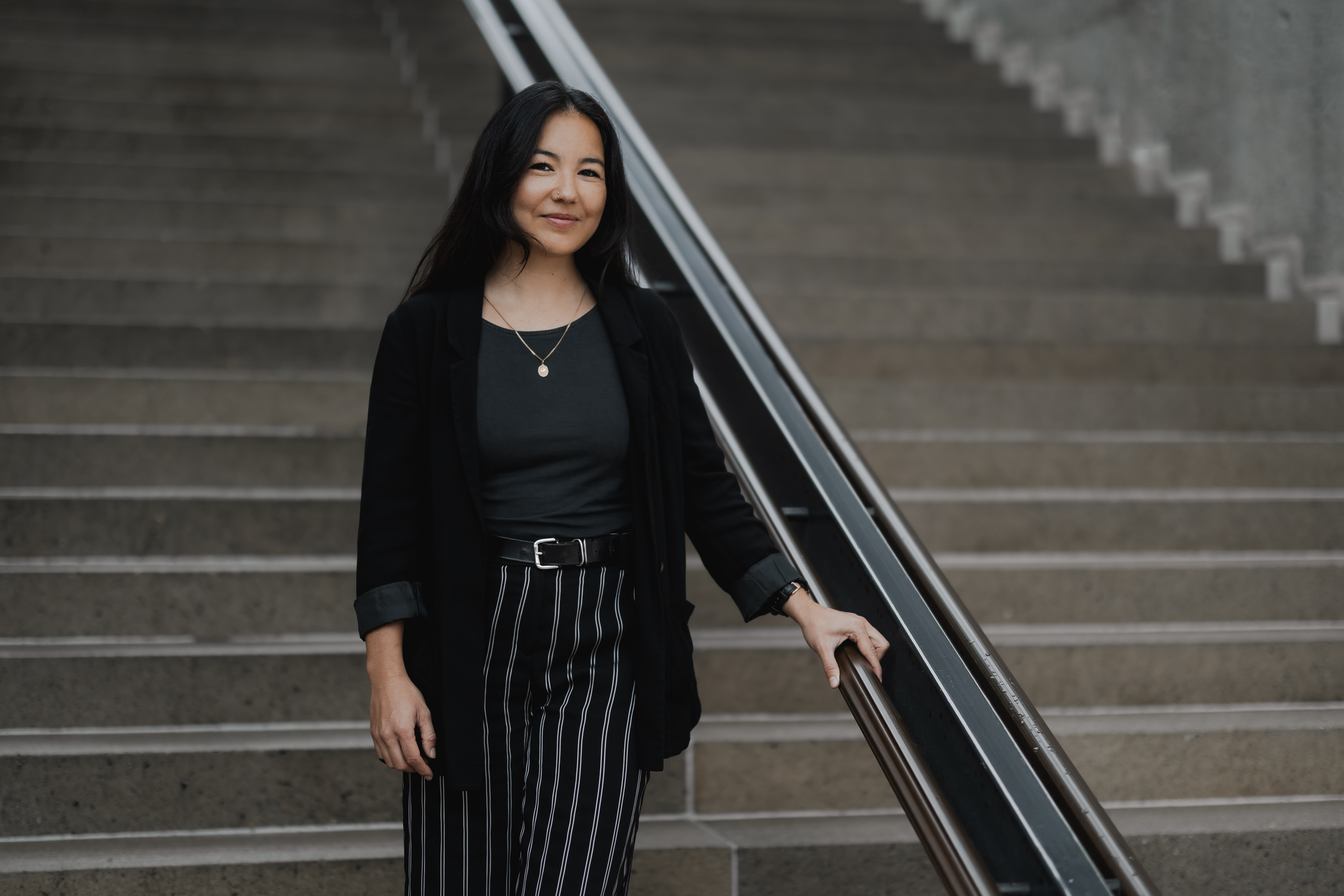 Eine Frau steht selbstbewusst auf einer Treppe. Sie trägt eine schwarze Jacke und gestreifte Hose und hat eine Hand auf dem Geländer. Sie lächelt in die Kamera und verkörpert das perfekte Lebenslauf-Porträt. Die Betonstufen im Hintergrund verleihen diesem Businessporträt eine professionelle Note.