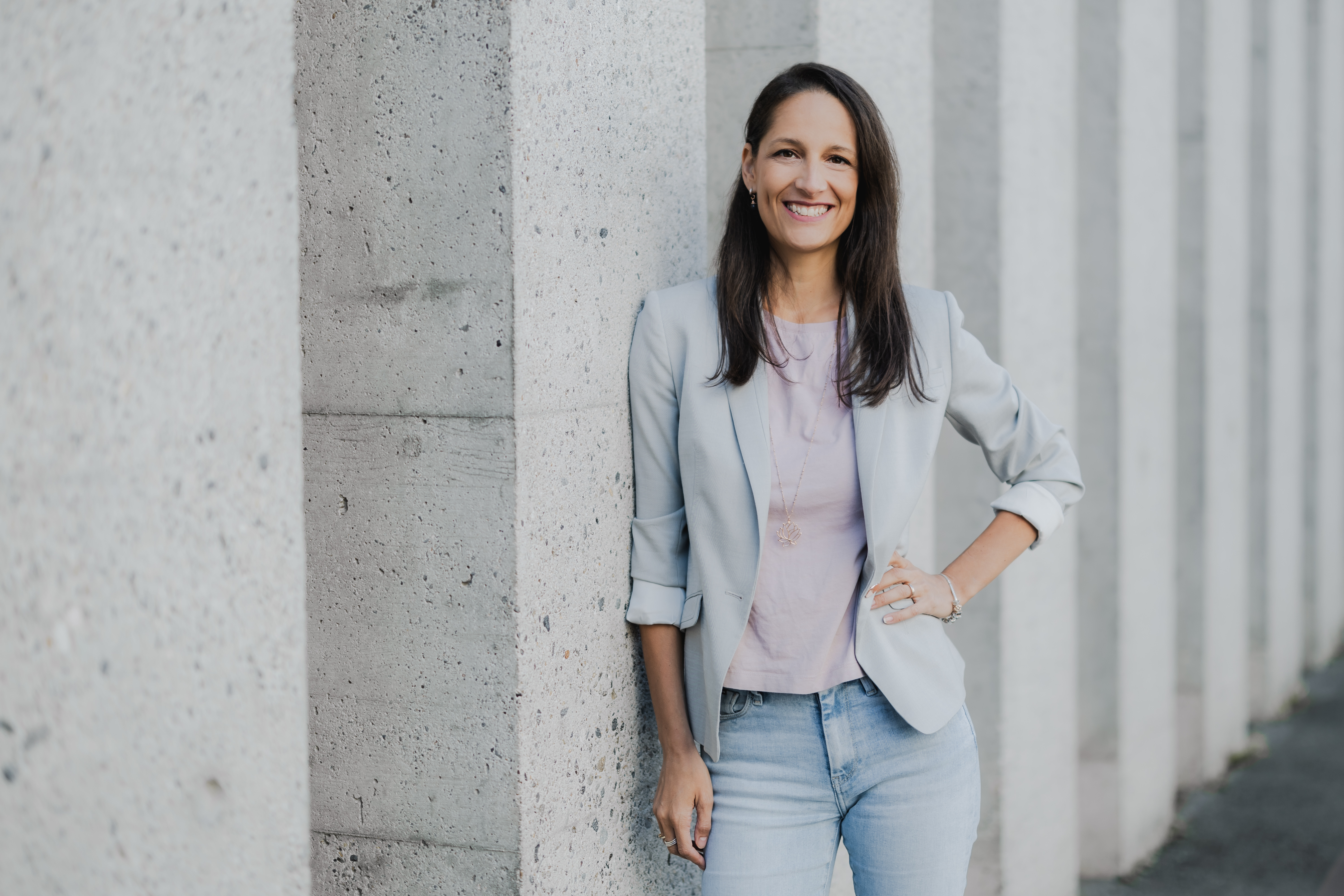 Eine Frau mit langen dunklen Haaren steht lächelnd vor einer Betonsäule und verkörpert das perfekte Businessporträt. Sie trägt einen hellgrauen Blazer, ein rosa Oberteil und Jeans. Im Hintergrund ist eine Reihe ähnlicher Säulen zu sehen, die eine ideale Kulisse für ein Lebenslauffoto bilden.