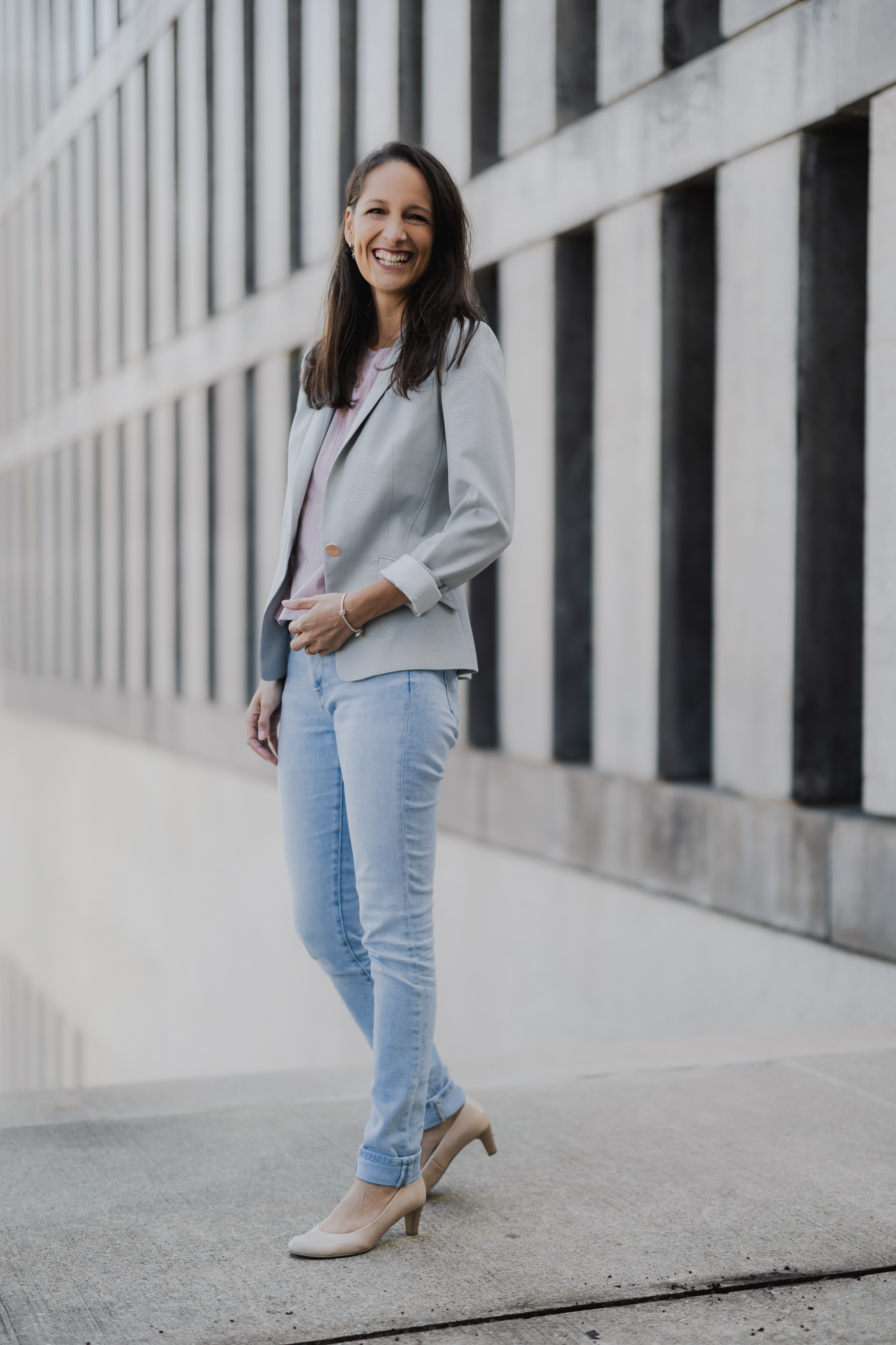 Eine Person mit langen dunklen Haaren lächelt und trägt einen hellgrauen Blazer, ein rosa Oberteil, hellblaue Jeans und beige Pumps. Sie steht auf einem Bürgersteig in der Stadt vor einem modernen Gebäude mit vertikalen Säulen im Hintergrund – ein ideales Business-Porträt oder Lebenslauffoto, das Selbstvertrauen und Stil ausstrahlt.