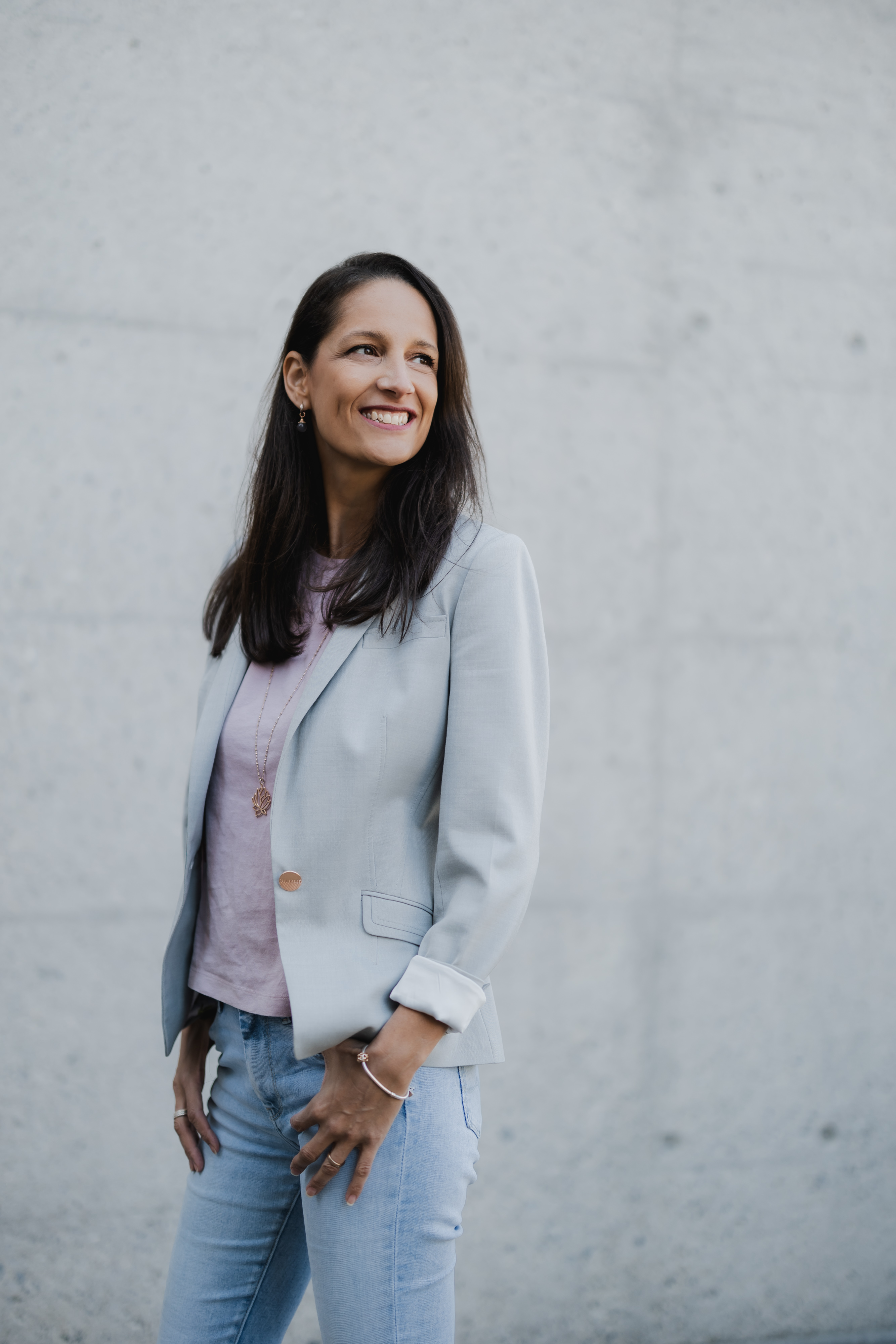Eine Frau mit langen dunklen Haaren lächelt selbstbewusst vor einer schlichten Betonwand und fängt damit die Essenz eines professionellen Porträts ein. Sie trägt einen hellgrauen Blazer, ein rosa Oberteil und blaue Jeans, ihre Hände ruhen auf ihren Hüften – perfekt für ein Lebenslauf- oder Bewerbungsfoto.