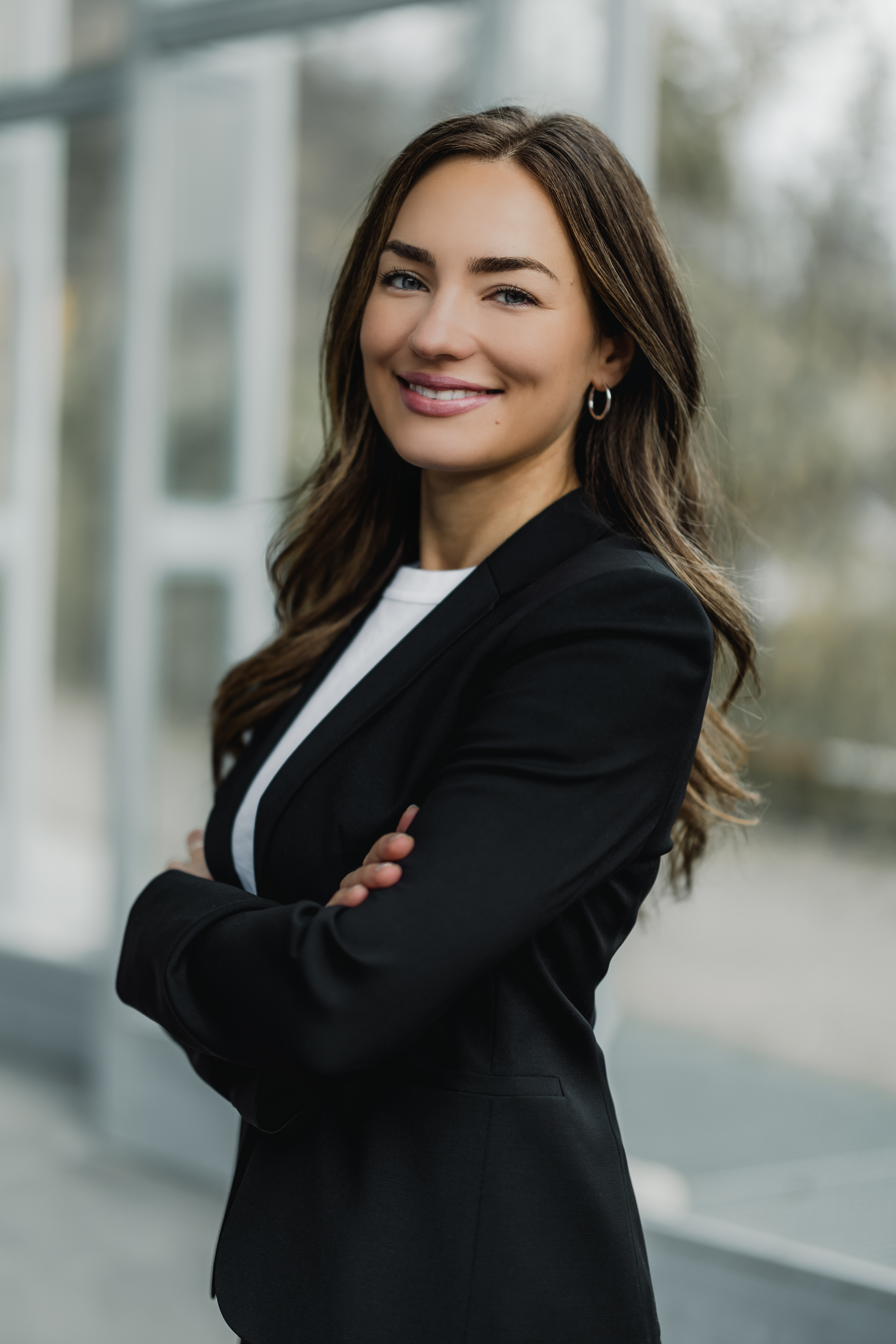 Eine Frau mit langen braunen Haaren steht selbstbewusst mit verschränkten Armen da. Sie trägt einen schwarzen Blazer über einem weißen Oberteil und lächelt leicht. Der Hintergrund ist leicht verschwommen und suggeriert eine helle Innenumgebung.
