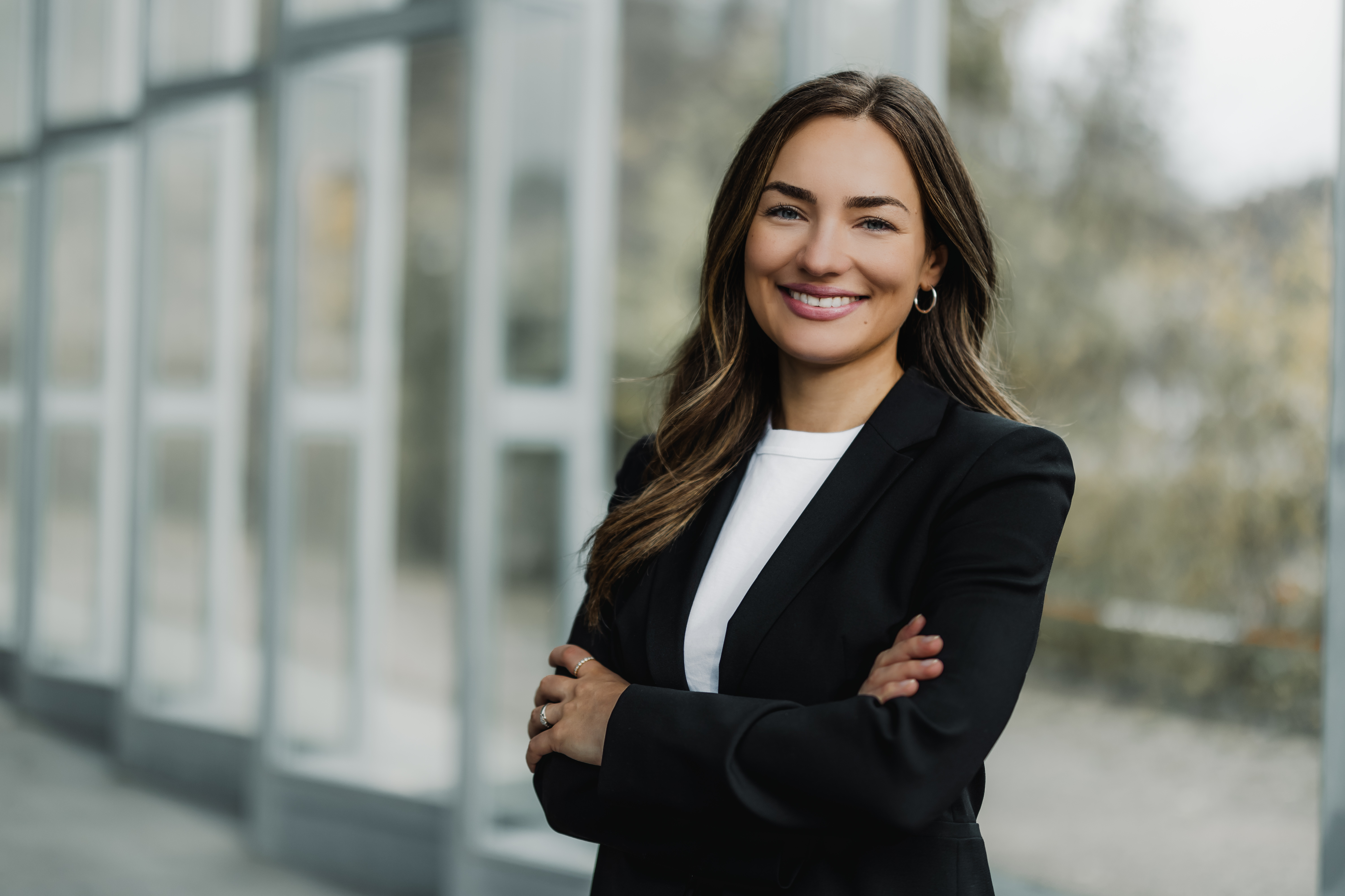 Eine Frau mit langen braunen Haaren und einem breiten Lächeln steht selbstbewusst mit verschränkten Armen da. Sie trägt einen schwarzen Blazer und ein weißes Hemd, vor einem verschwommenen Hintergrund mit Glasfenstern.