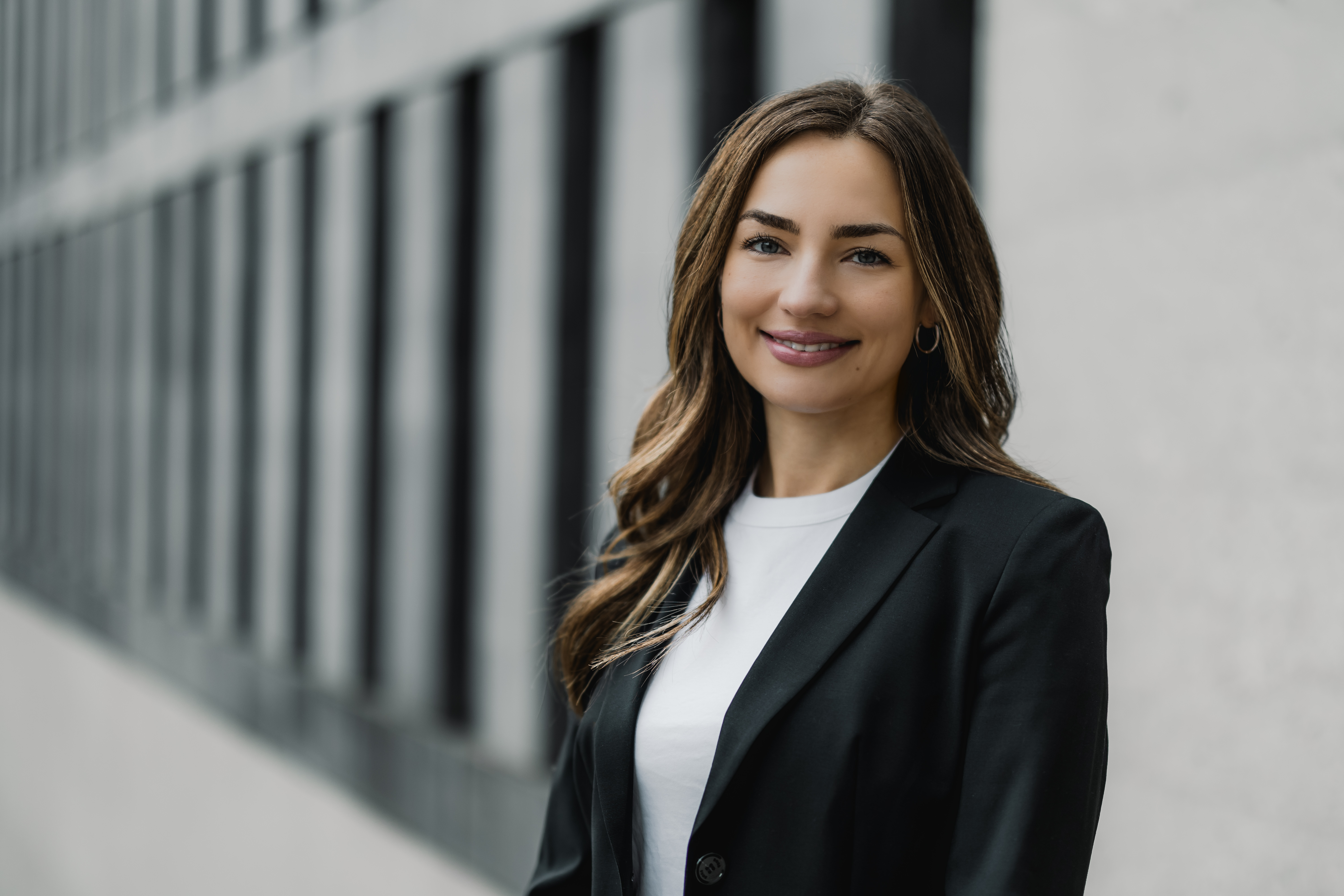 Eine Frau mit langen braunen Haaren lächelt und trägt einen schwarzen Blazer über einem weißen Hemd. Sie steht draußen vor einem Gebäude mit vielen Fenstern.
