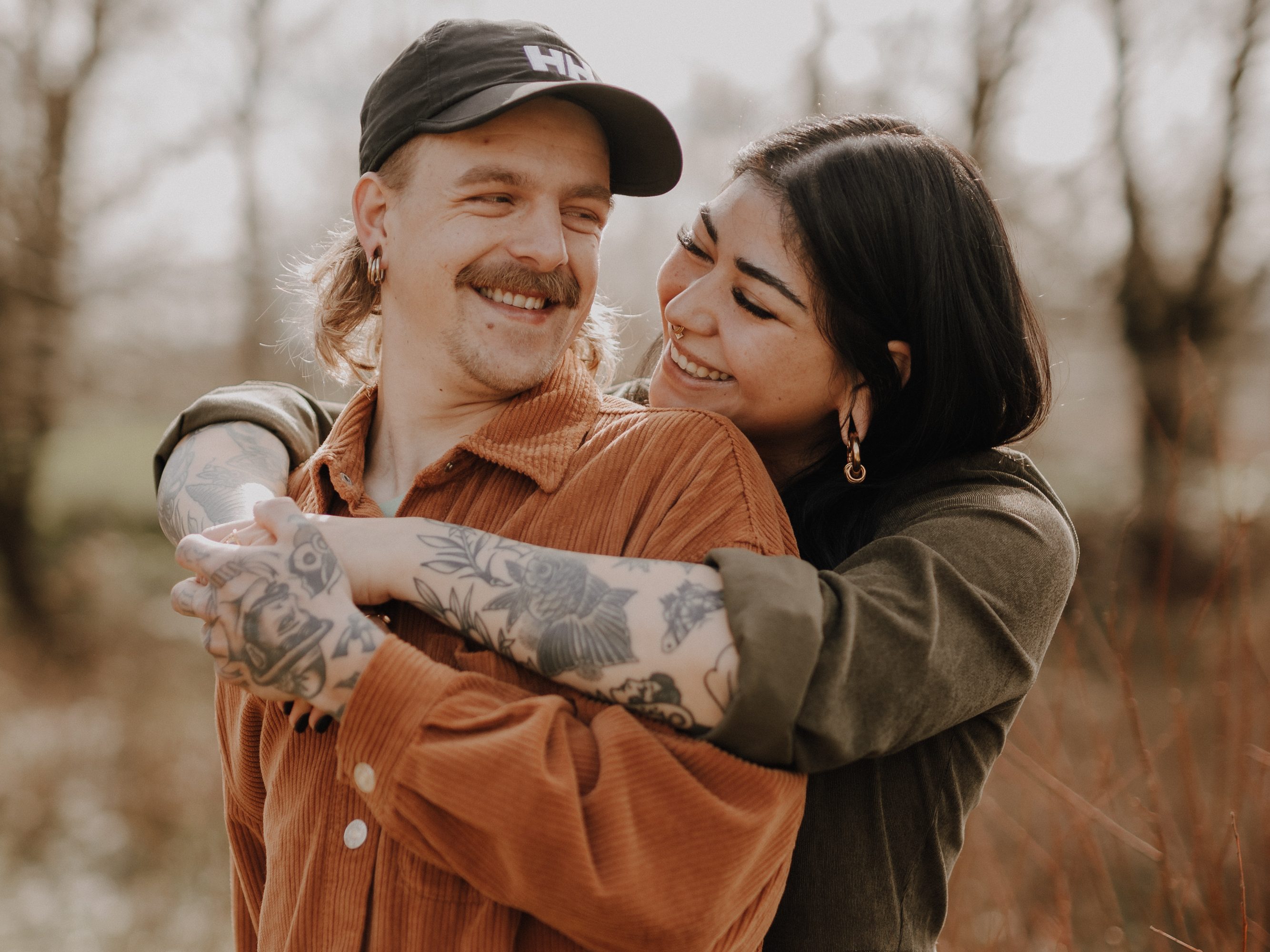 Eine lächelnde Frau mit langen dunklen Haaren umarmt einen lächelnden Mann mit Schnurrbart und Tattoos, der eine Mütze und ein braunes Hemd trägt, von hinten. Sie stehen während ihres Paarfotografie-Fotoshootings im Freien zwischen kahlen Bäumen und trockenem Laub. Die Frau wirkt freudig, als sie den Mann ansieht.