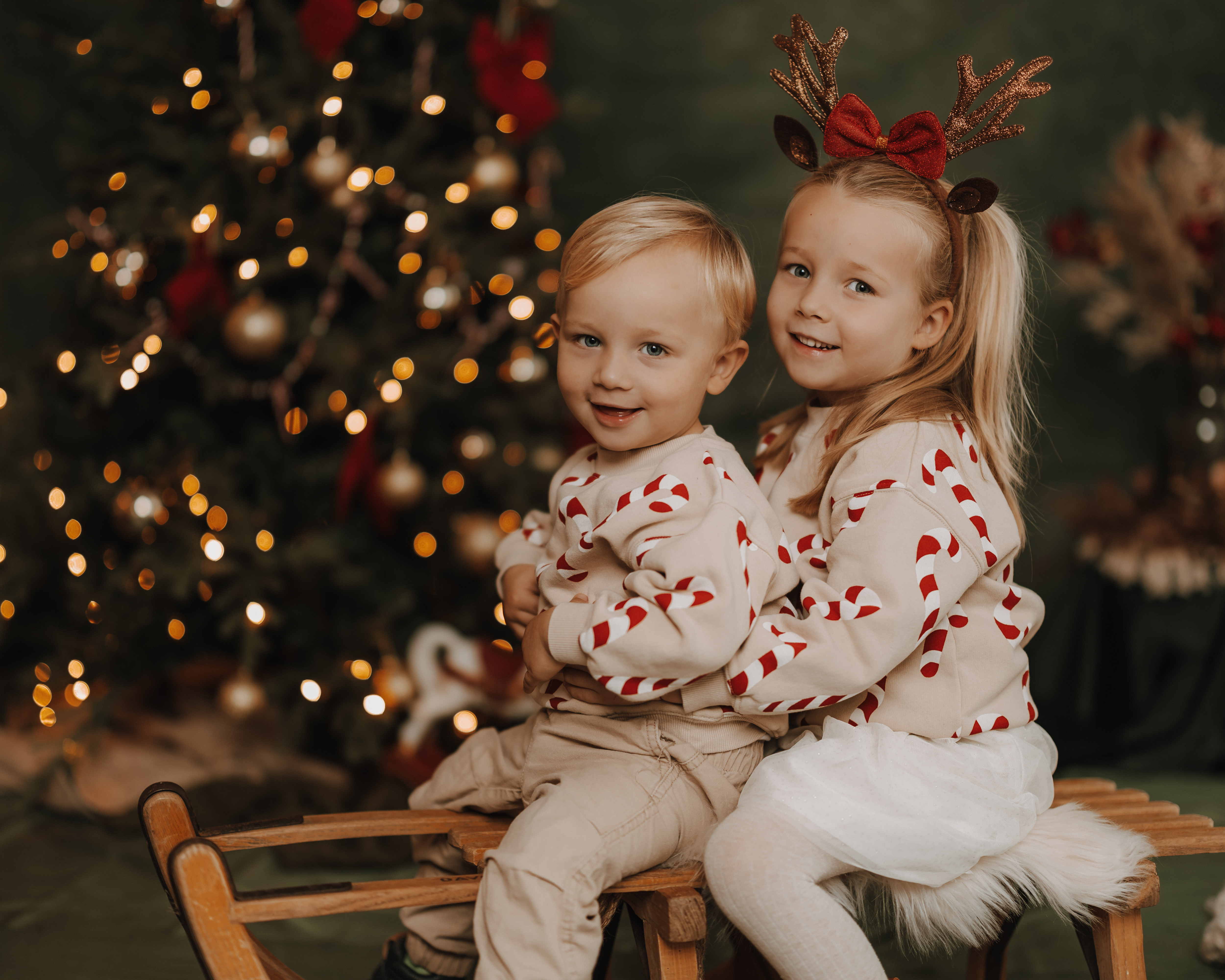 Zwei kleine Kinder sitzen auf einem Holzschlitten vor einem geschmückten Weihnachtsbaum. Beide tragen passende beige Pullover mit roten Zuckerstangen. Das Mädchen mit dem Rentier-Stirnband umarmt den Jungen von hinten - perfekt für ein charmantes Weihnachtsfoto oder Weihnachtsshooting.