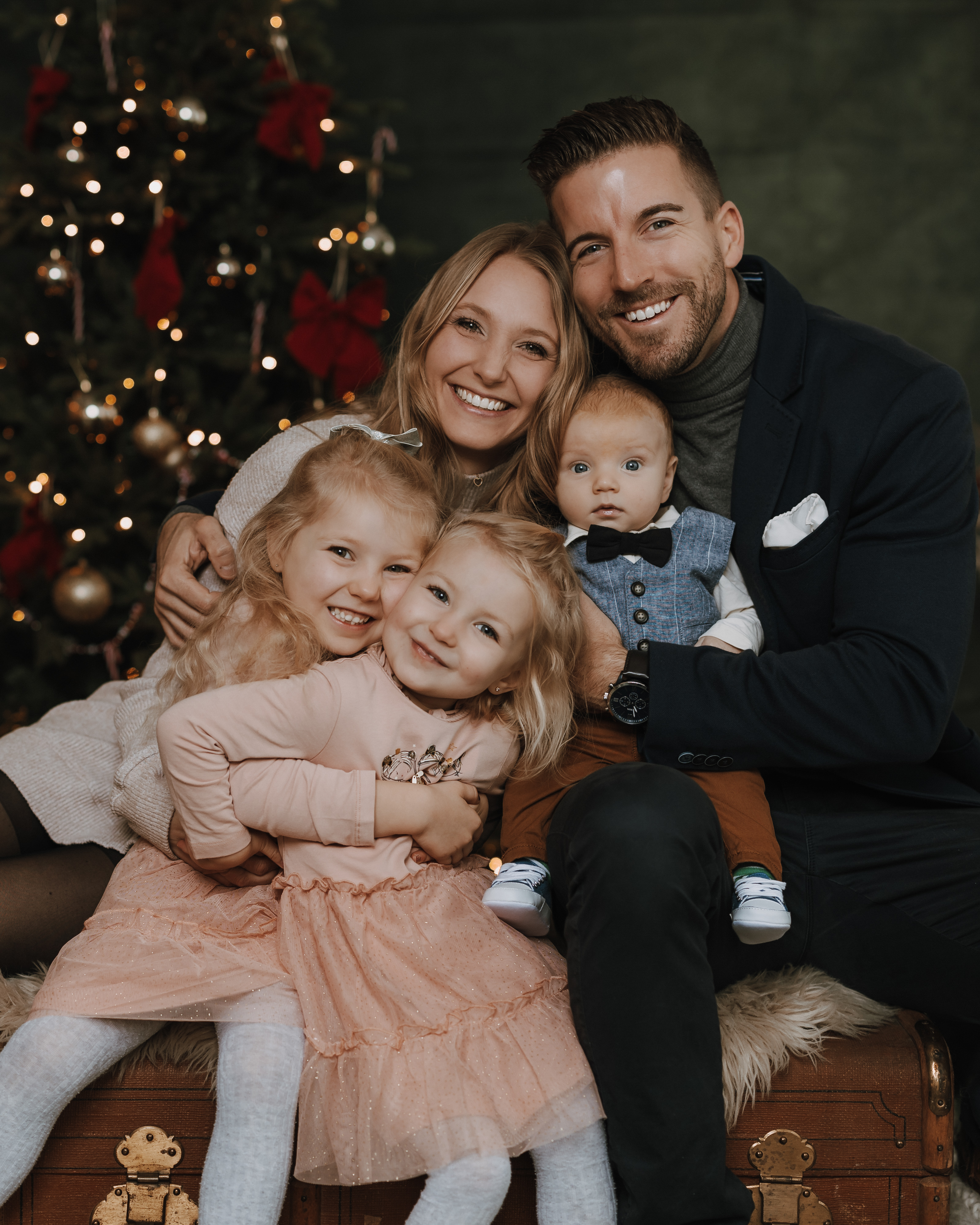 Eine lächelnde fünfköpfige Familie posiert für ein Weihnachts-Fotoshooting vor einem geschmückten Baum. Die Eltern sitzen hinter ihren drei kleinen Kindern, die festlich in Pastellfarben und mit einer Fliege gekleidet sind. Alle sehen glücklich und bereit für das Weihnachtsshooting aus.
