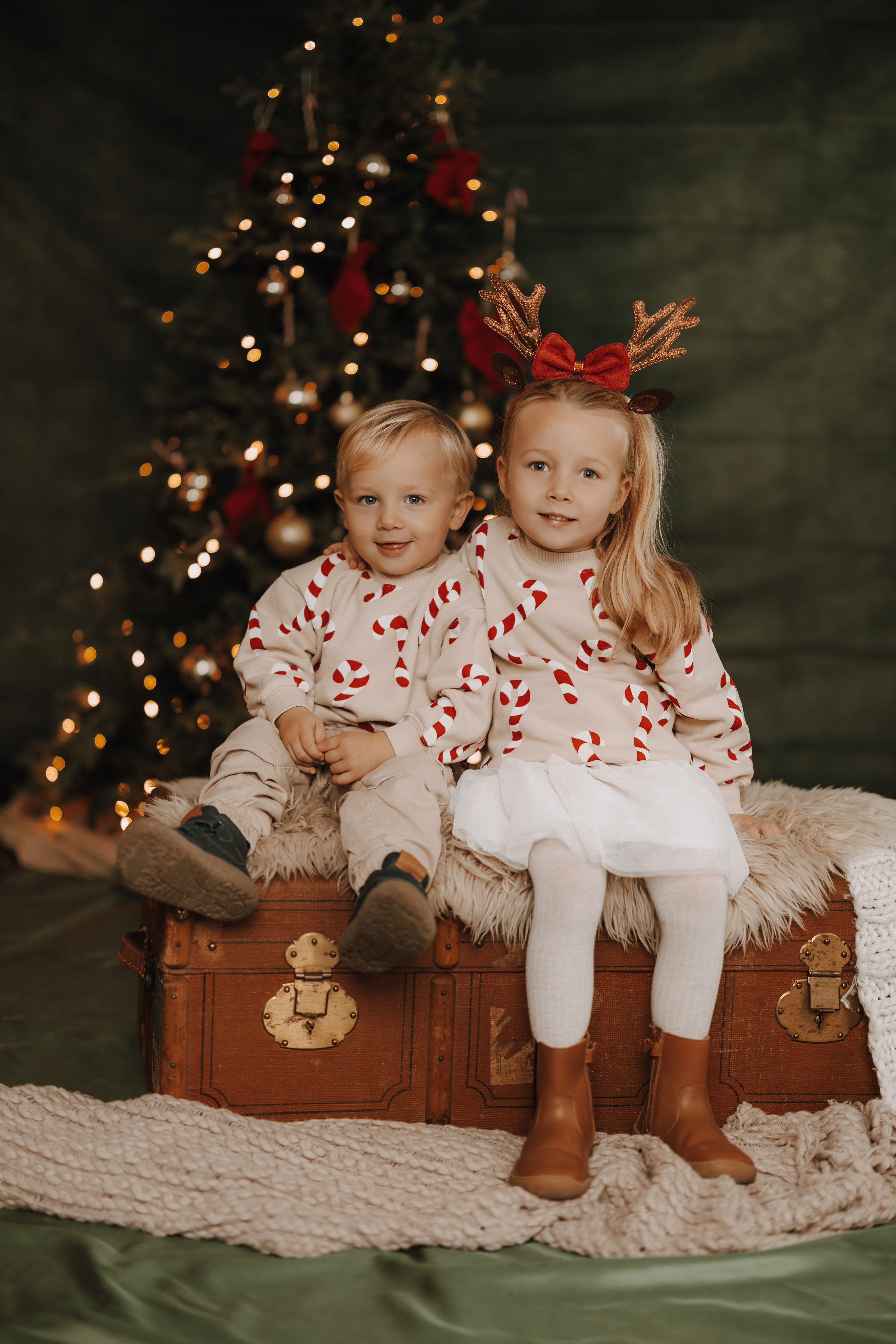 Zwei kleine Kinder in passenden festlichen Outfits sitzen auf einem alten Baumstamm vor einem geschmückten Weihnachtsbaum; das Mädchen trägt ein Rentiergeweih, und beide lächeln warm in die Kamera - perfekt für ein unvergessliches Weihnachtsfoto.