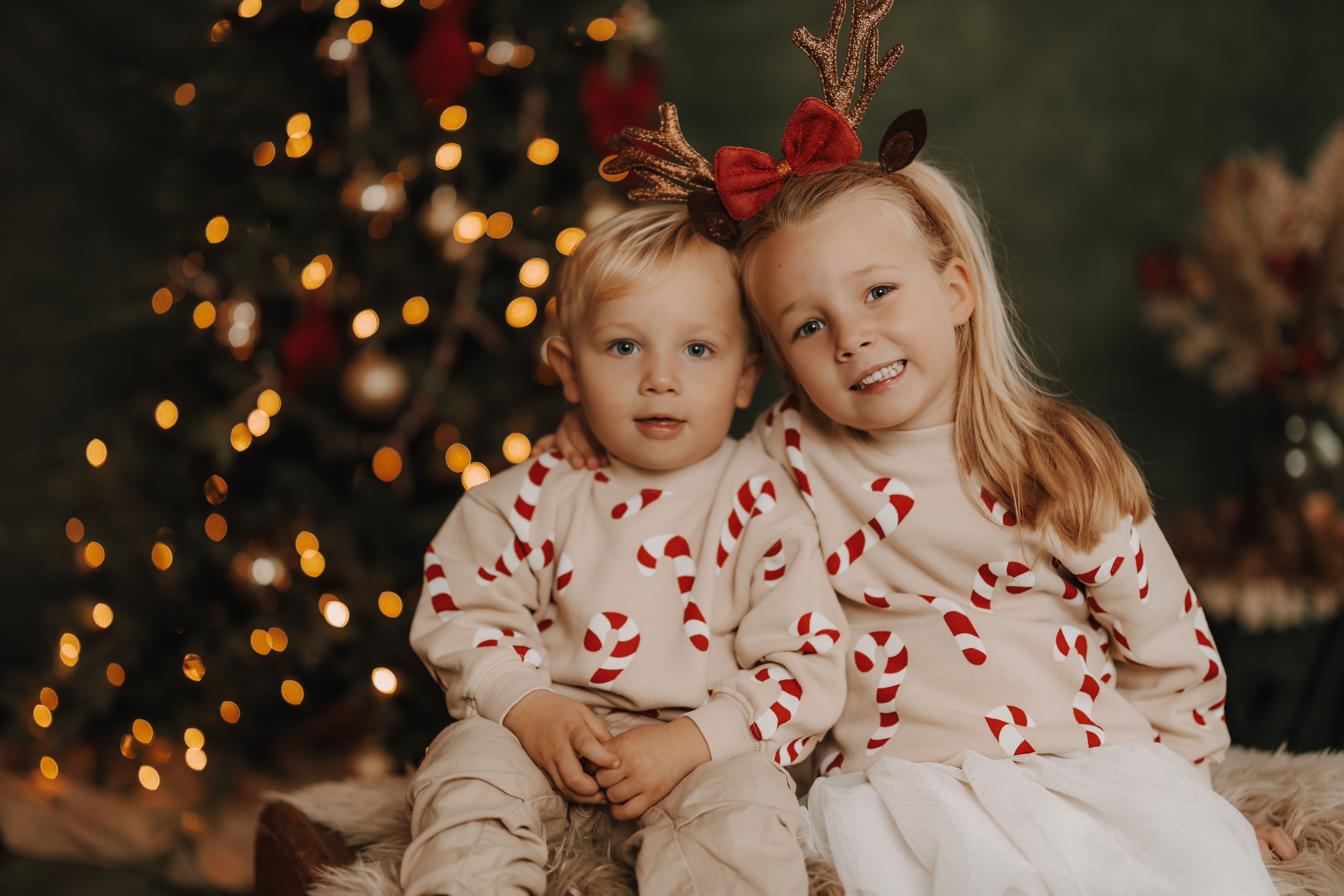 Zwei kleine Kinder sitzen während eines gemütlichen Weihnachts-Fotoshootings zusammen vor einem Weihnachtsbaum mit Lichtern. Beide tragen passende cremefarbene Hemden mit roten Zuckerstangen, und das ältere Kind mit dem Rentiergeweih umarmt das jüngere und lächelt in die Kamera.